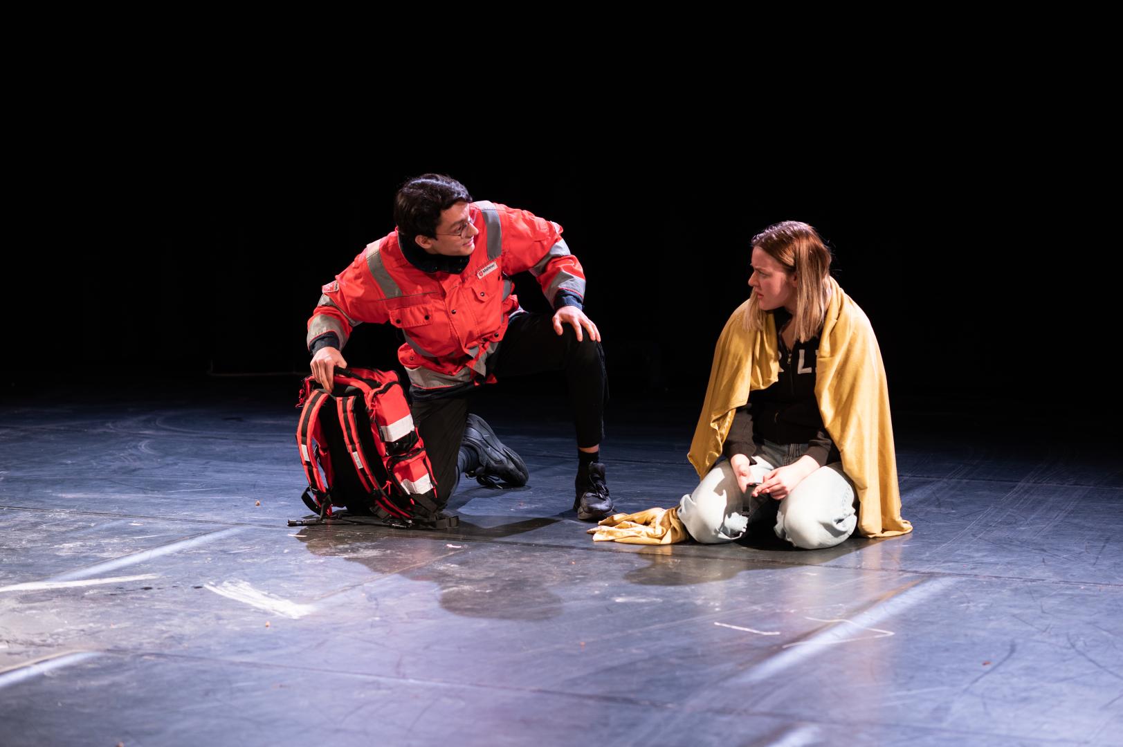 Schauspieler in Sanitäterkostüm kniet sich zu einer Schauspielerin mit einem Handtuch um die Schultern herunter.