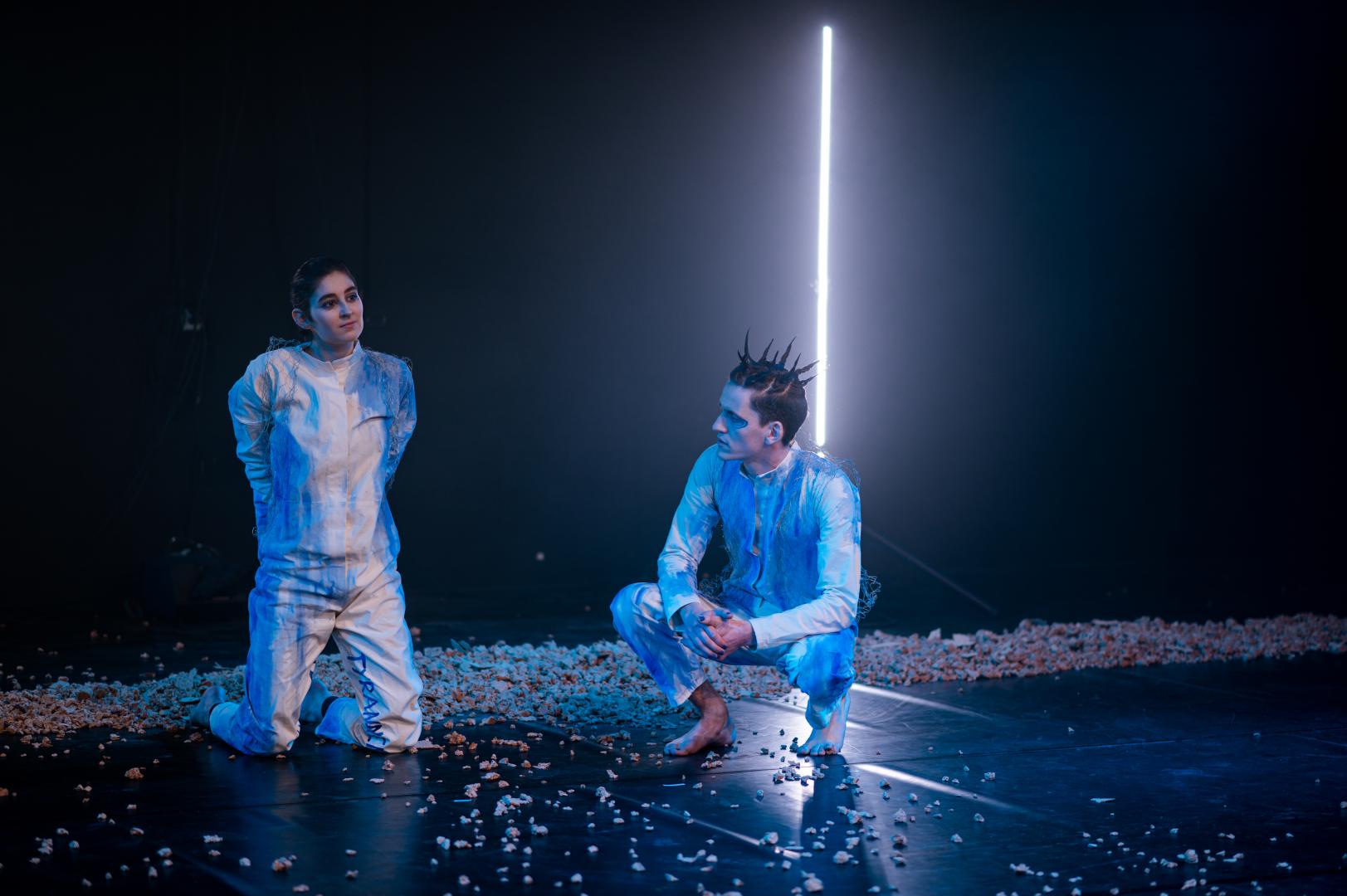 Ein Schauspieler und eine Schauspielerin in blauem Bühnenlicht auf dem Boden sitzend.