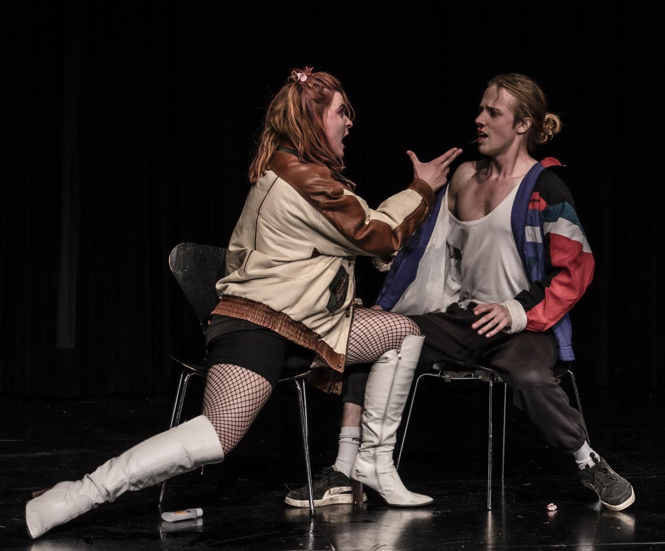 Schauspielerin zeigt mit ihrer Hand, die zu einer Pistole geformt ist, auf einen Schauspieler, der aus der Nase blutet.