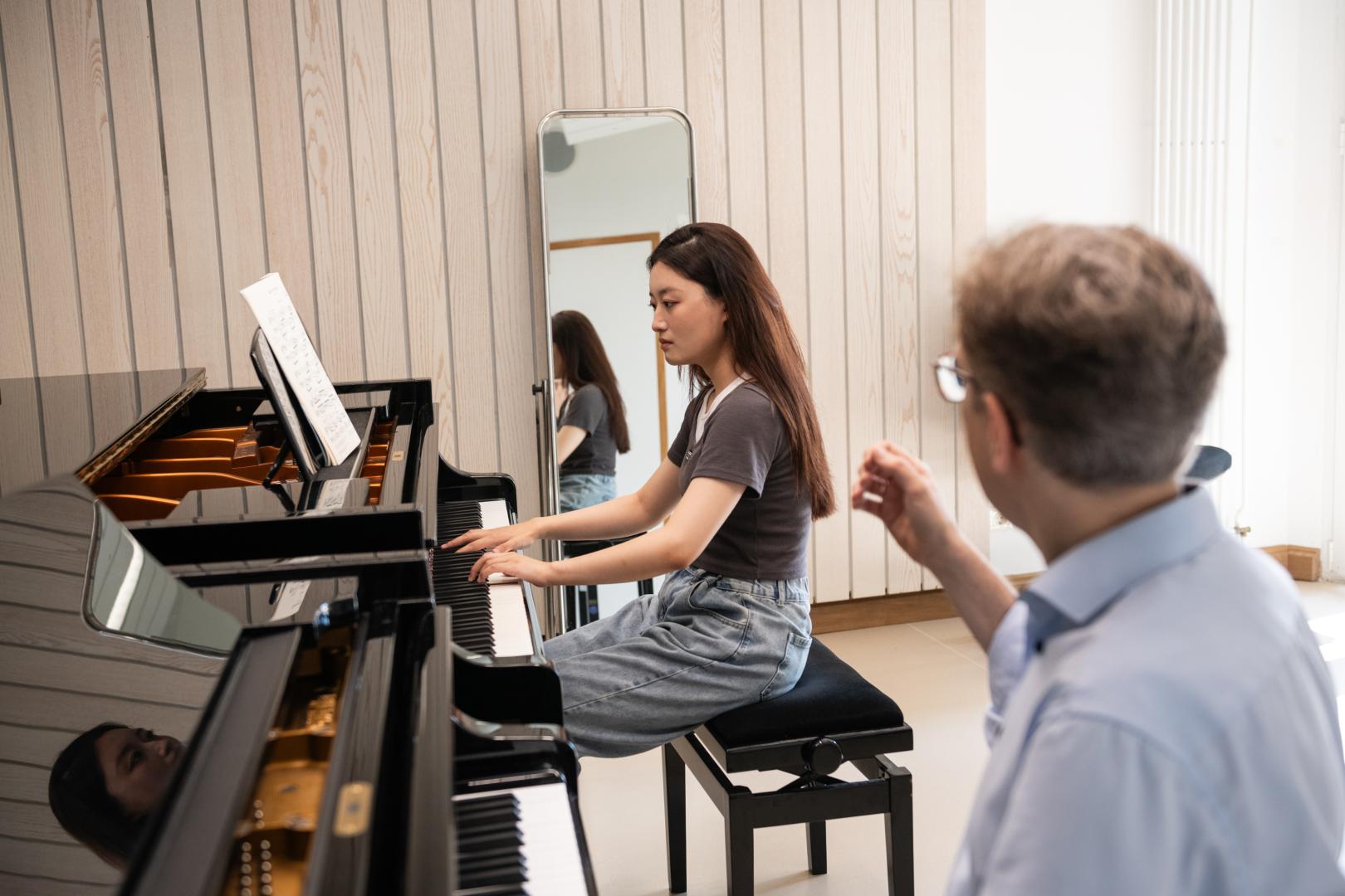 Klavier-Einzelunterricht: Eine Studentin spielt am Flügel, im Vordergrund sieht man den Professor, der ihr währenddessen etwas erklärt. 