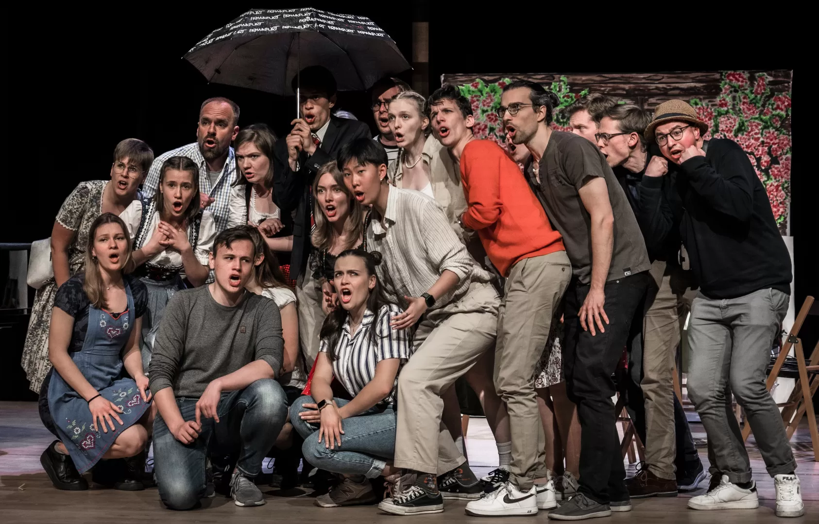 Große Gurppe von Darsteller*innen im Weißen Rössl auf der Bühne, eng aneinander unter einem einzigen kleinen Regenschirm.