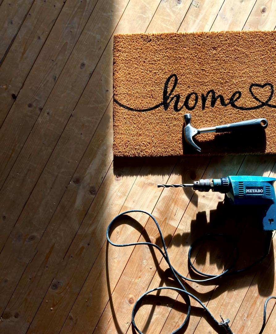 Aus der Vogelperspektive sieht man einen Holzboden, auf diesem rechts an der Seite eine Fußmatte mit dem Wort "Home", darunter rechts ein blauer Elektrobohrer mit Strohkabel.
