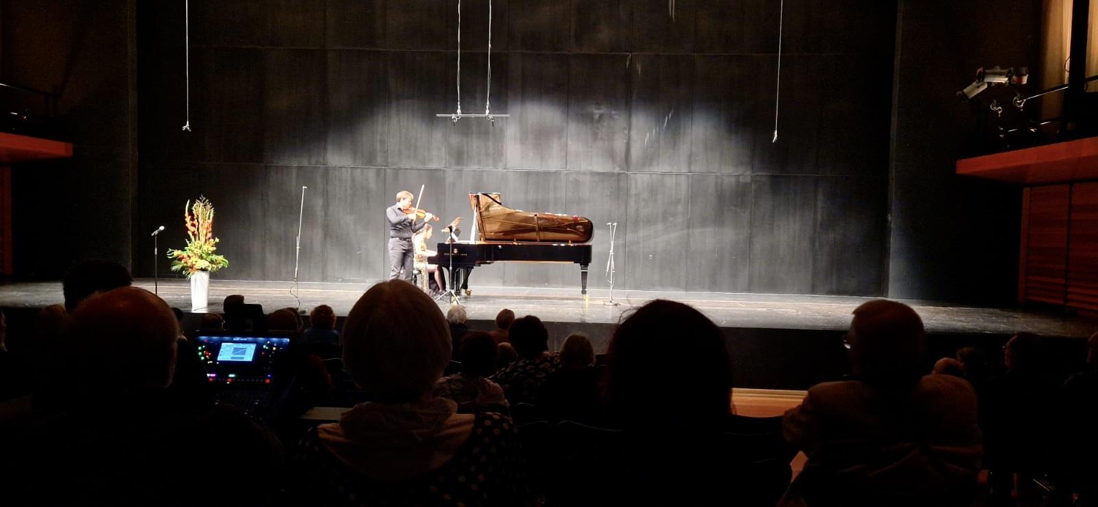 Geiger und Pianistin auf der erleuchteten Bühne in einem dunklen Saal mit Publikum. 