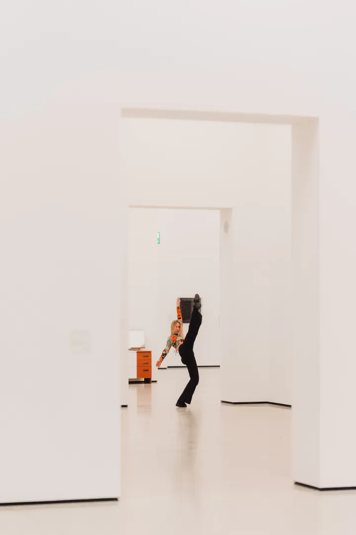 In der Mitte des Bildes posiert eine Tänzerin in einem Durchgang, umgeben von weißen Wänden. Im Hintergrund ist eine Kommode und ein schwarzes Bild zu sehen. Die Person trägt eine lange, schwarze Hose und ein buntes langärmliges Oberteil. Der Blick ist zur Kamera gerichtet.   