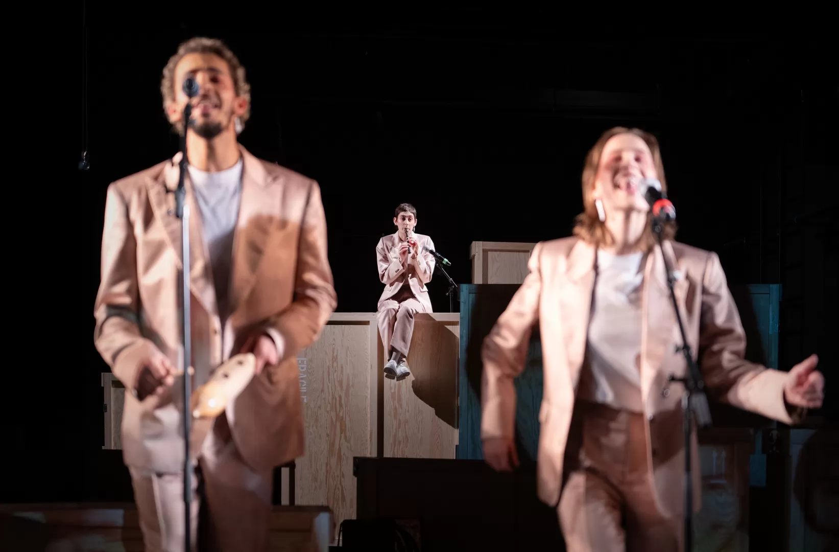 Zwei Personen singen im Vordergrund, im Hintergrund sitzt eine Person und spielt Flöte.