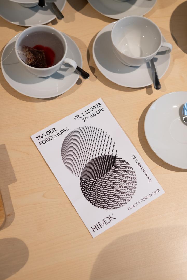 Auf einem Tisch mit vollen und leeren Tassen liegt ein geschlossenes Programmheft, auf dem das Datum, der Zeitraum und der Ort des Tags der Forschung erkennbar werden. Die Grafik zeigt zwei Kreise mit unterschiedlichen schwarz-weißen Mustern, die sich überlagern. In der Schnittfläche der beiden Kreise zeigt sich ein neues Muster, das nicht aus der Überlagerung der beiden ursprünglichen Muster hervorgeht.