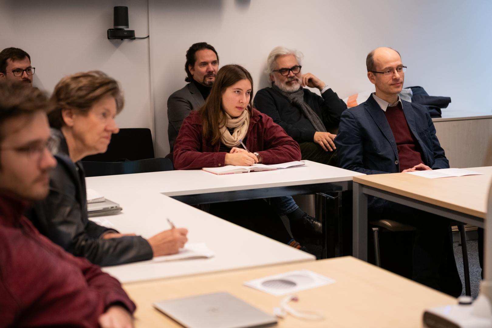 Eine Gruppe von sieben Zuhörenden sitzt in einem Vorlesungssaal an Tischen. Sie richten die Aufmerksamkeit nach rechts vorne, aber die präsentierende Person selbst ist nicht im Bild.