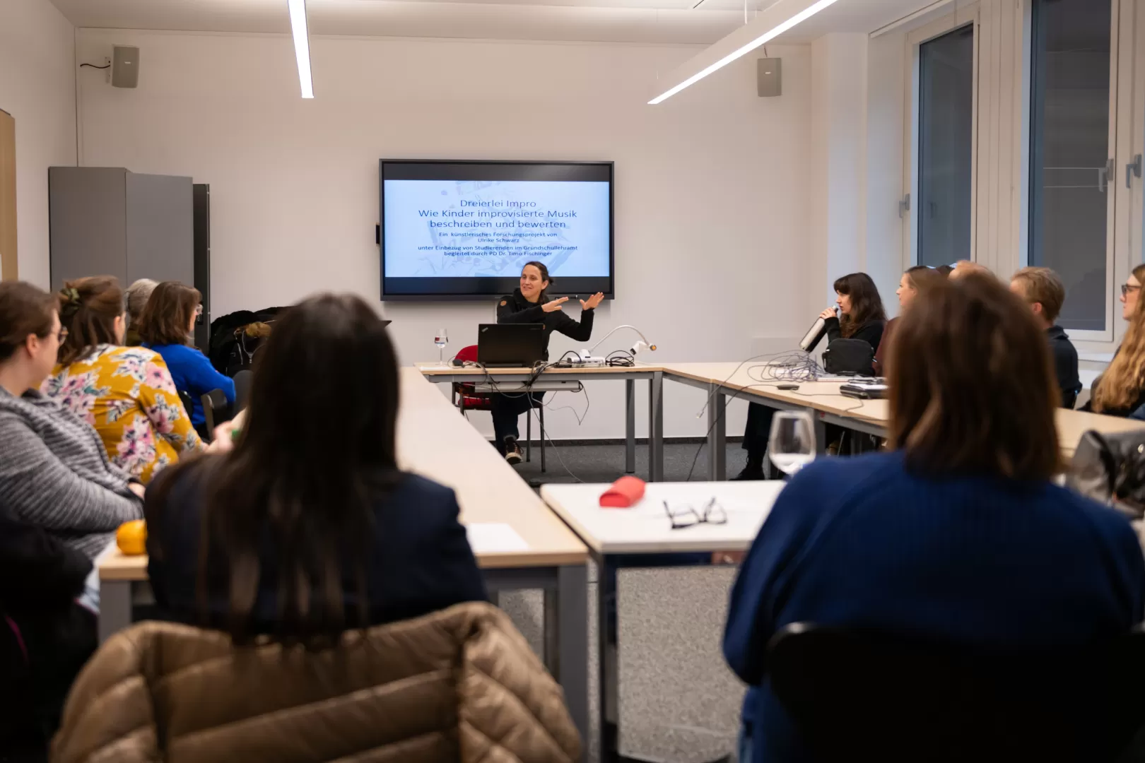 Im Vorder- und im Mittelgrund ist eine Gruppe von mehreren Zuhörenden sitzt an Tischen im Kreis. Fokussiert im Zentrum des Bildes ist eine Vortragende, die im Hintergrund an einem Tisch sitzt und mit den Händen eine erklärende Geste ausführt. Hinter der eine Präsentation auf einem Bildschirm erkennbar wird: Dort ist zu lesen: „Dreierlei Impro – Wie Kinder improvisierte Musik beschreiben und bewerten“.