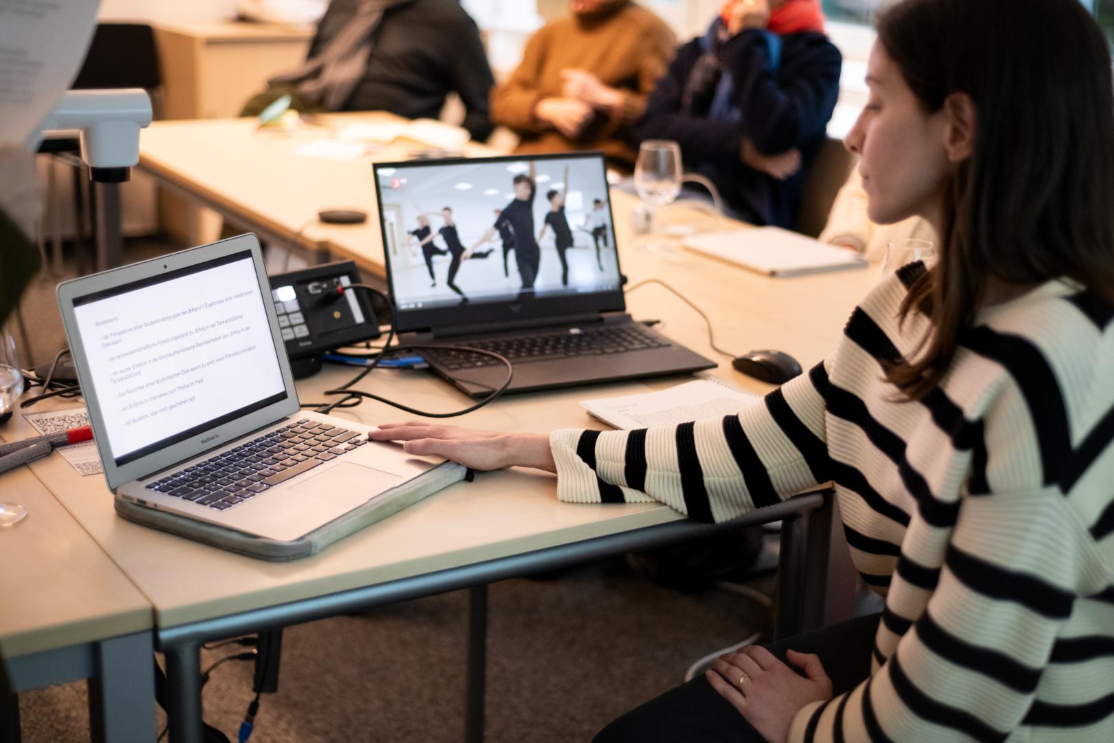 Im Vordergrund rechts sehen wir eine Frau im Profil, die sitzt und auf zwei Laptops links von ihr blickt. Ein Laptop zeigt Text, der andere das Still eines Tanzvideos. Im Hintergrund sind unscharf Zuhörende zu erkennen.