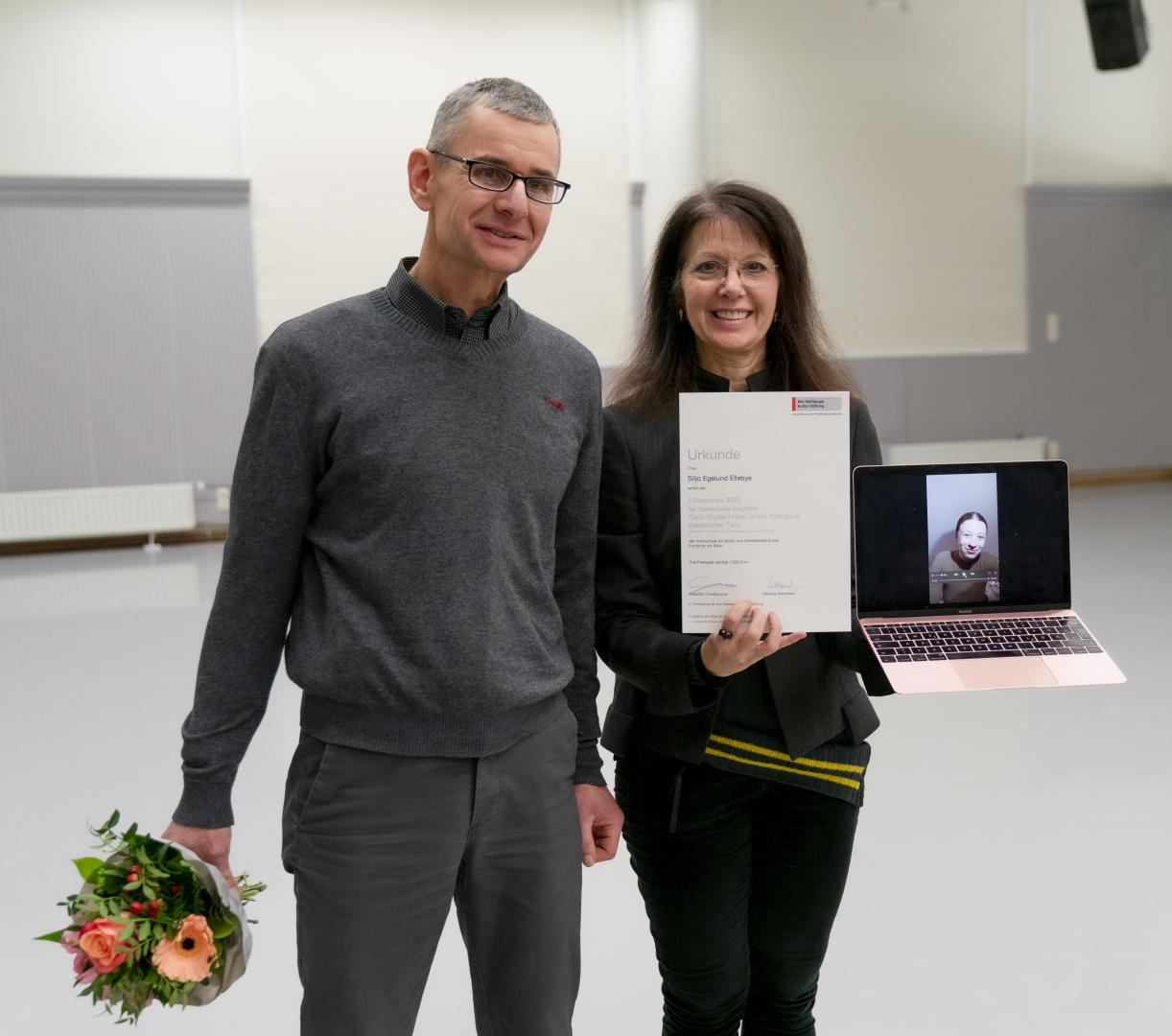 Silja Egelund Ellebye ist per Laptop zugeschaltet, um ihren Förderpreis entgegenzunehmen. Ein Mann und eine Frau halten Blumenstrauß, Urkunde und Laptop.