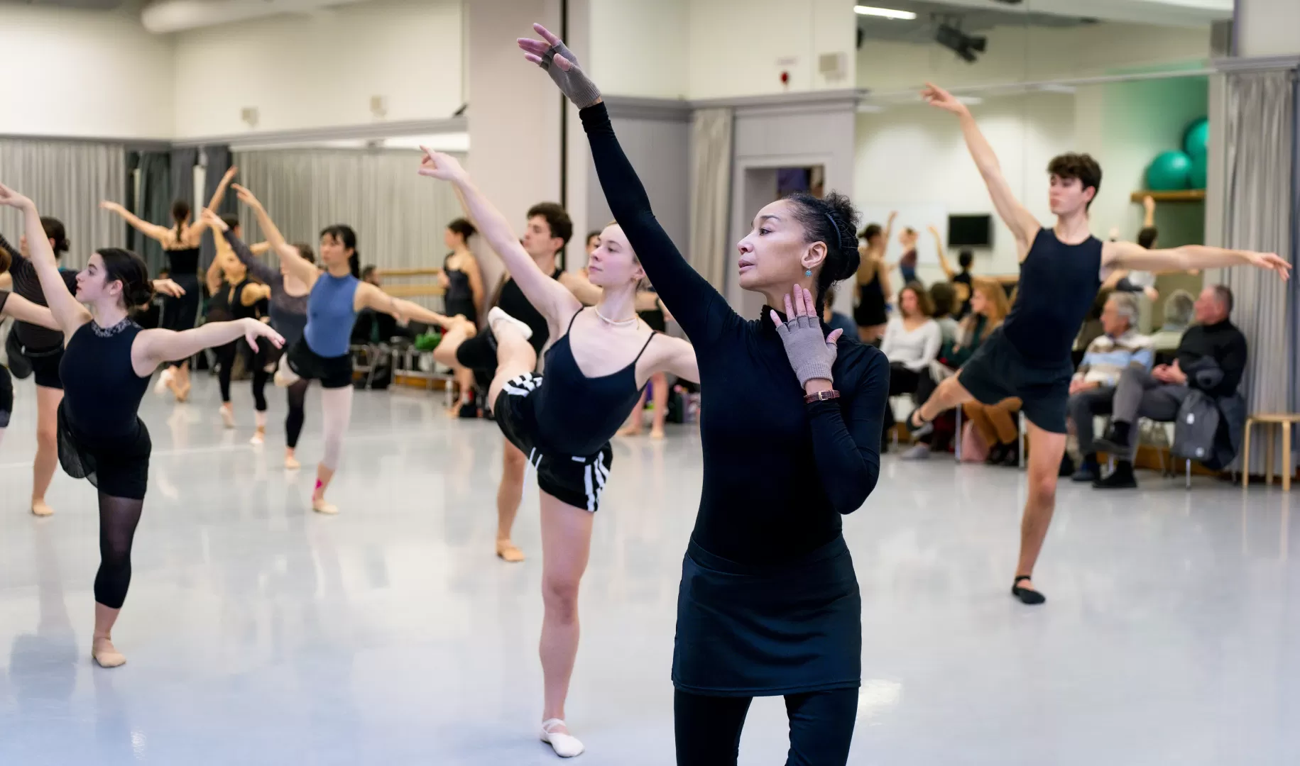 Die Ballettlehrerin streckt ihren rechten Arm elegant nach oben. Hinter ihr tanzen ihre Schüler:innen.
