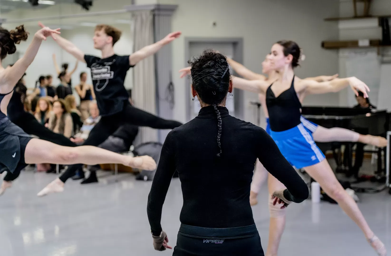 Die Ballettlehrerin von hinten. Unscharf dahinter üben Tänzer:innen Sprünge (grand jetés).