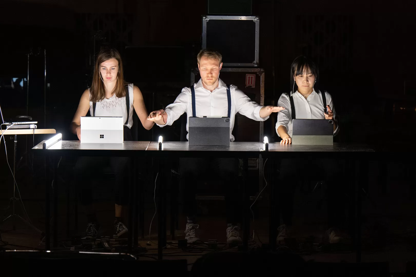 Drei Performer*innen im dunklen Raum, vor ihnen Laptops auf dem Tisch.