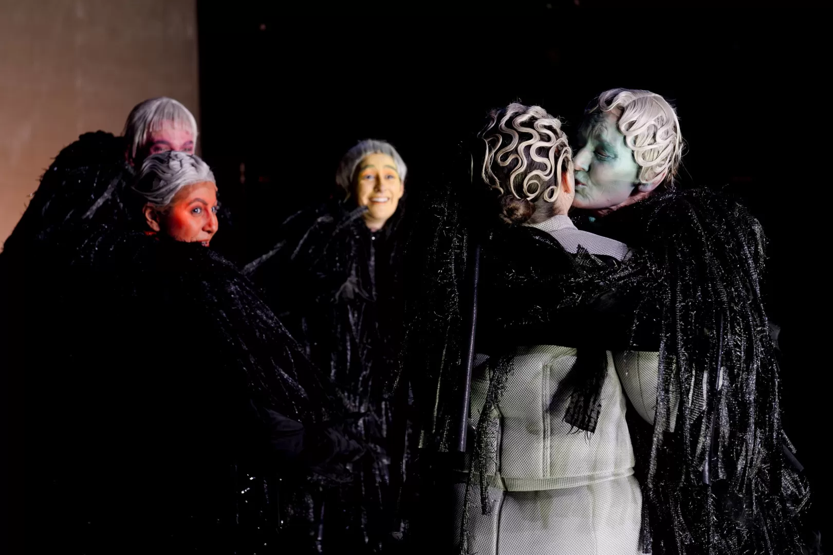 Fünf schwarz gekleidete Personen mit bunten Gesichtern, zwei sich küssend, die anderen schauen zu ihnen hin.