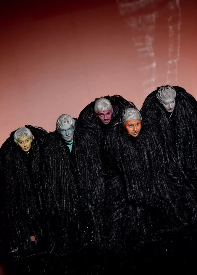 Fünf schwarz gekleidete Personen mit bunten Gesichtern und in gebückter Haltung.