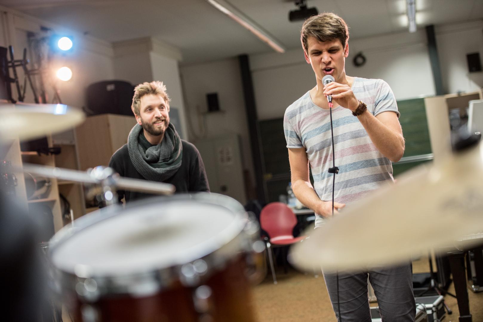 Ein Band-Coaching: Hinter einem Schlagzeug schaut ein Coach lächeln einem Studenten beim Singen zu.