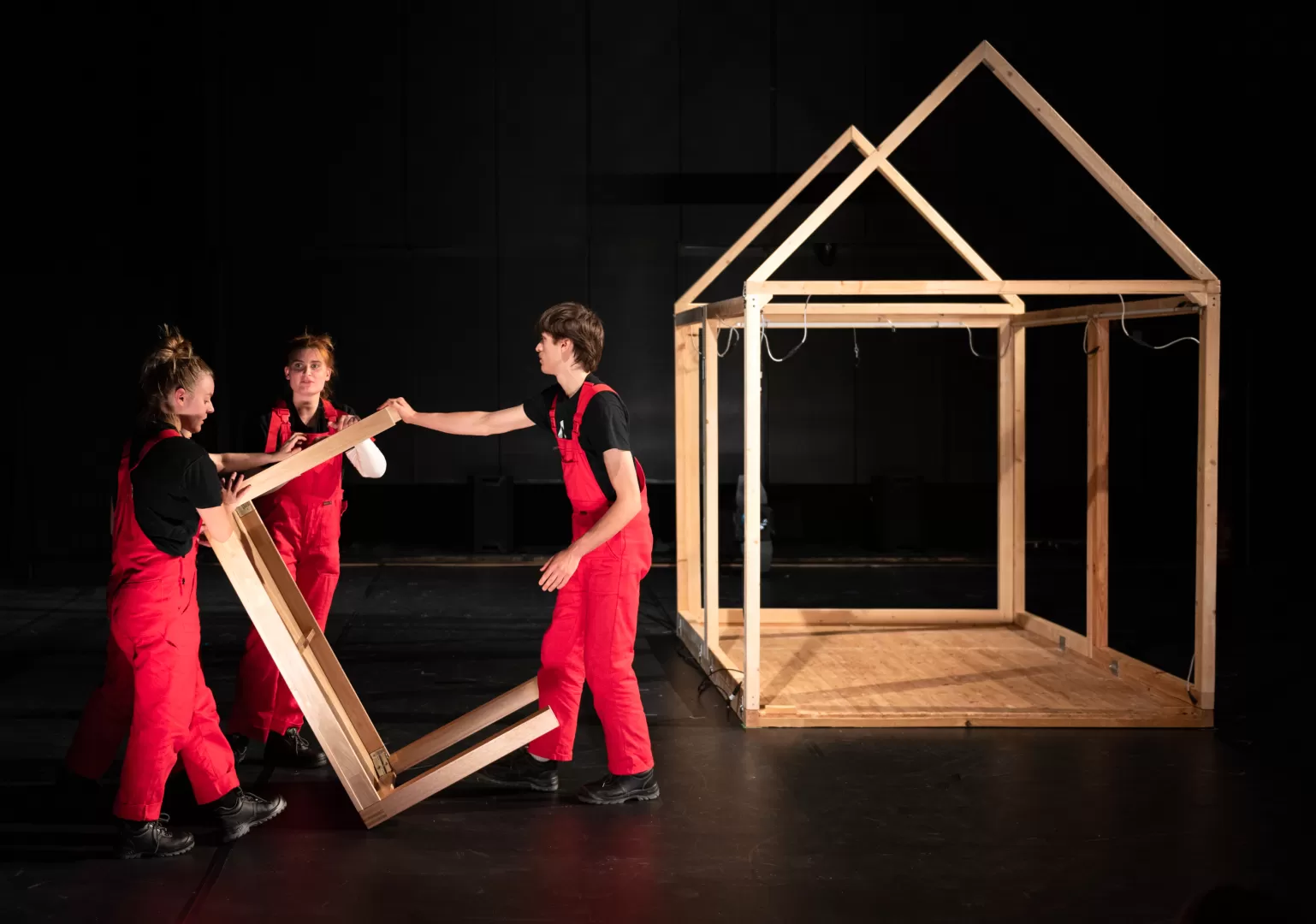 Drei Personen in roten Latzhosen tragen einen Holztisch.