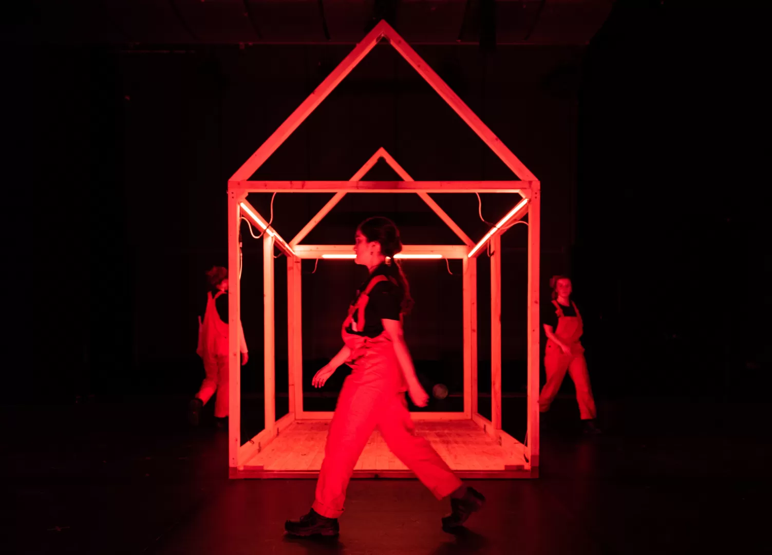 Drei Personen in roten Latzhosen laufen um das Gestell von einem Haus.