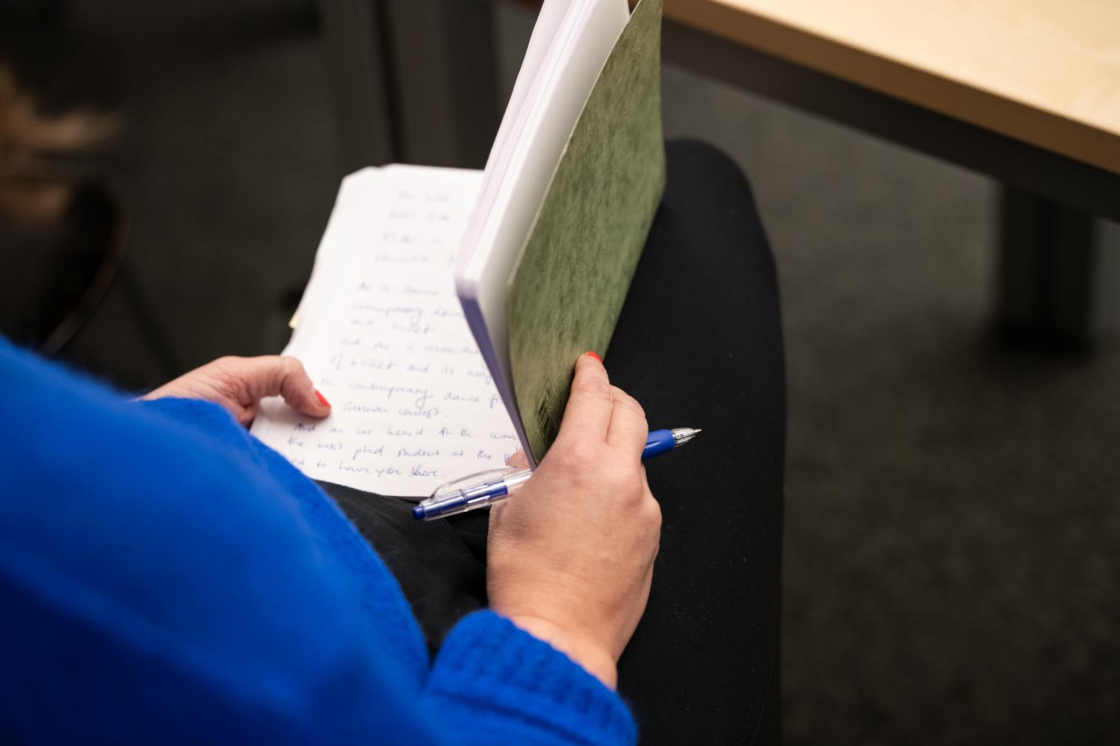 Wir sehen über die rechte Schulter einer sietzenden Person in blauem Pulli auf ein geöffnetes Notizheft und einen Kugelschreiber, die von der Person in Händen gehalten werden.