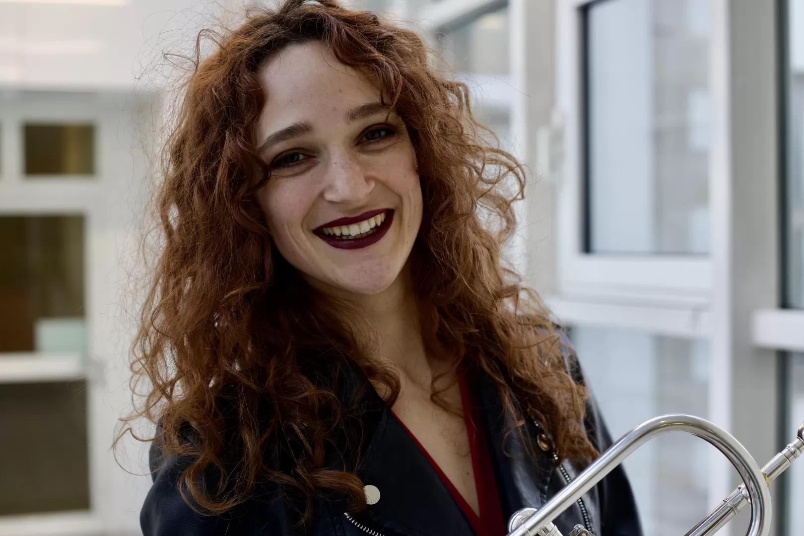 Portrait von Ines Hartmuth: Sie hat rotbraune Locken und lacht in die Kamera. Im Arm hält sie ihre Trompete.