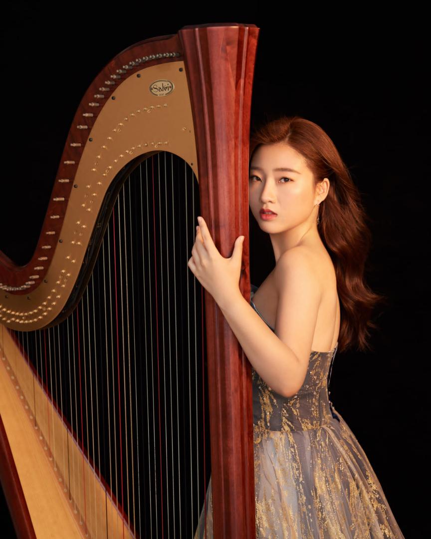 Harfe-Studentin Nan Wang, in einem edlen Kleid, hat die Hand und das Gesicht an der Harfe und blickt ernst in die Kamera.