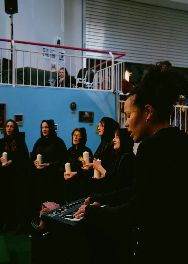 Eine Frau spielt und singt an einem Keyboard. Dahinter stehen die anderen Personen in schwarzen Kutten und mit Kerzen in der Hand.