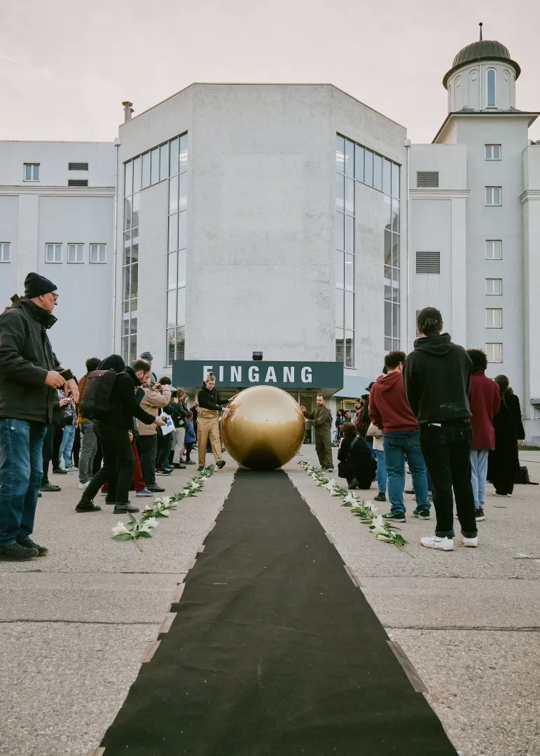 Zwei Männer rollen die goldene Kugel einen schwarzen langen Teppich herab. An den Seiten steht das Publikum und schaut zu.