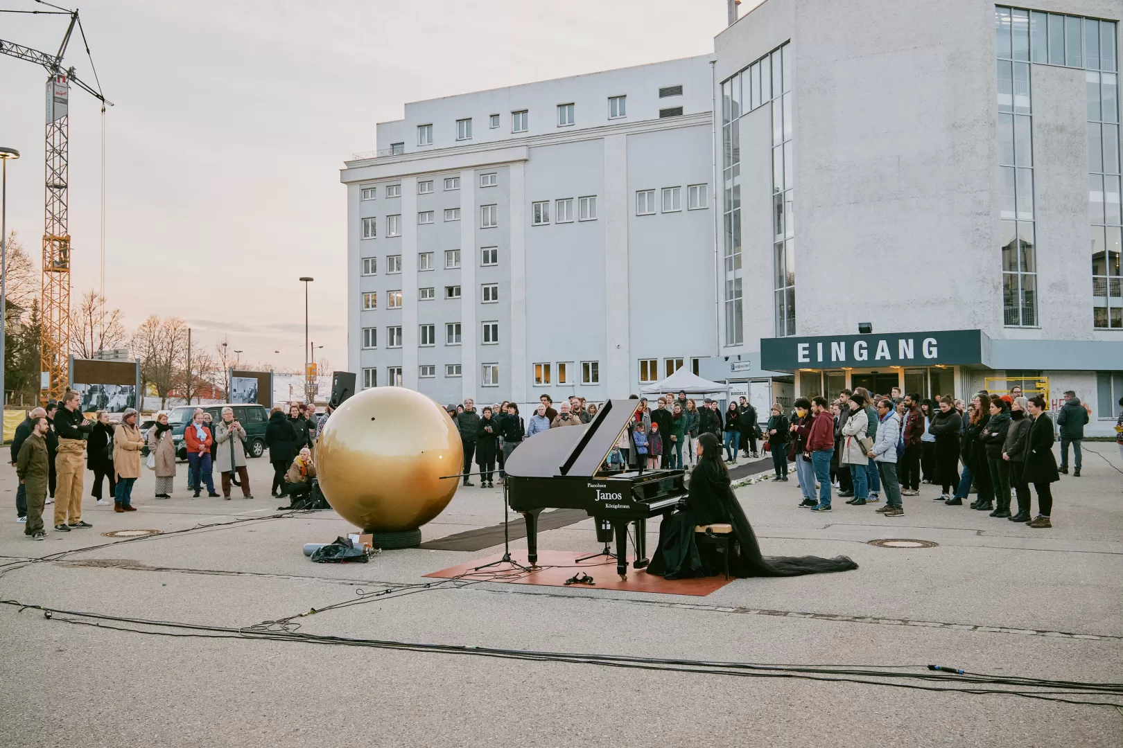 Auf einem Parkplatz hat sich eine Menschenmenge im Halbkreis um das Geschehen gestellt. Die goldene Kugel steht am Ende des schwarzen Teppichs. Daneben sitzt eine junge Frau mit schwarzem Umgang und Teufelshörnern an einem Flügel.