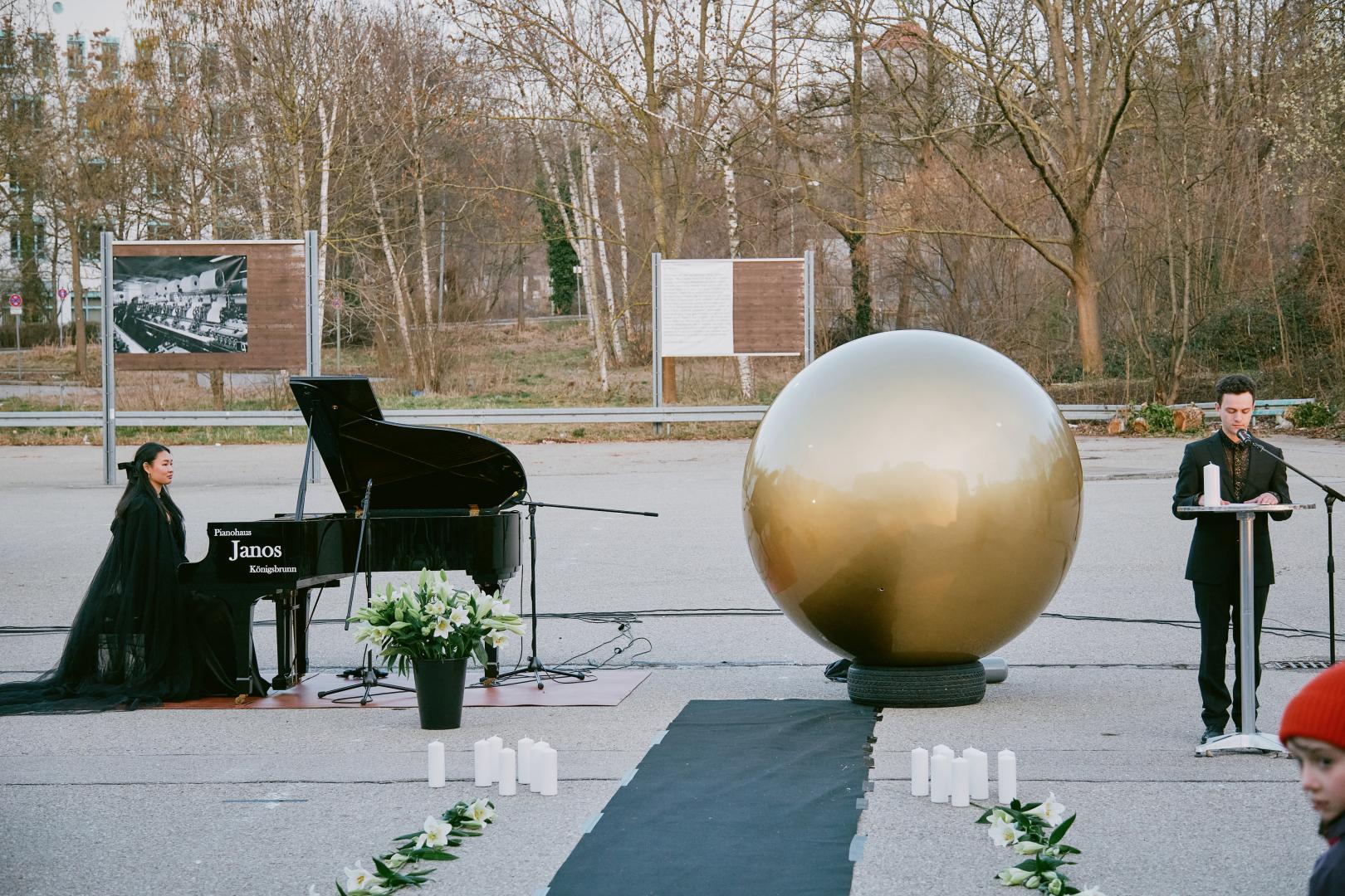 Links die Pianistin, in der Mitte die goldene Kugel, rechts ein Trauerredner an einem Pult.