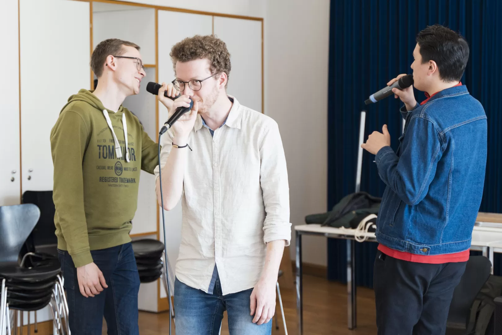 Drei Sänger mit Mikrofonen im Coaching. Die Person in der Mitte schaut nach vorne, die beiden äußeren singen sich gegenseitig an.