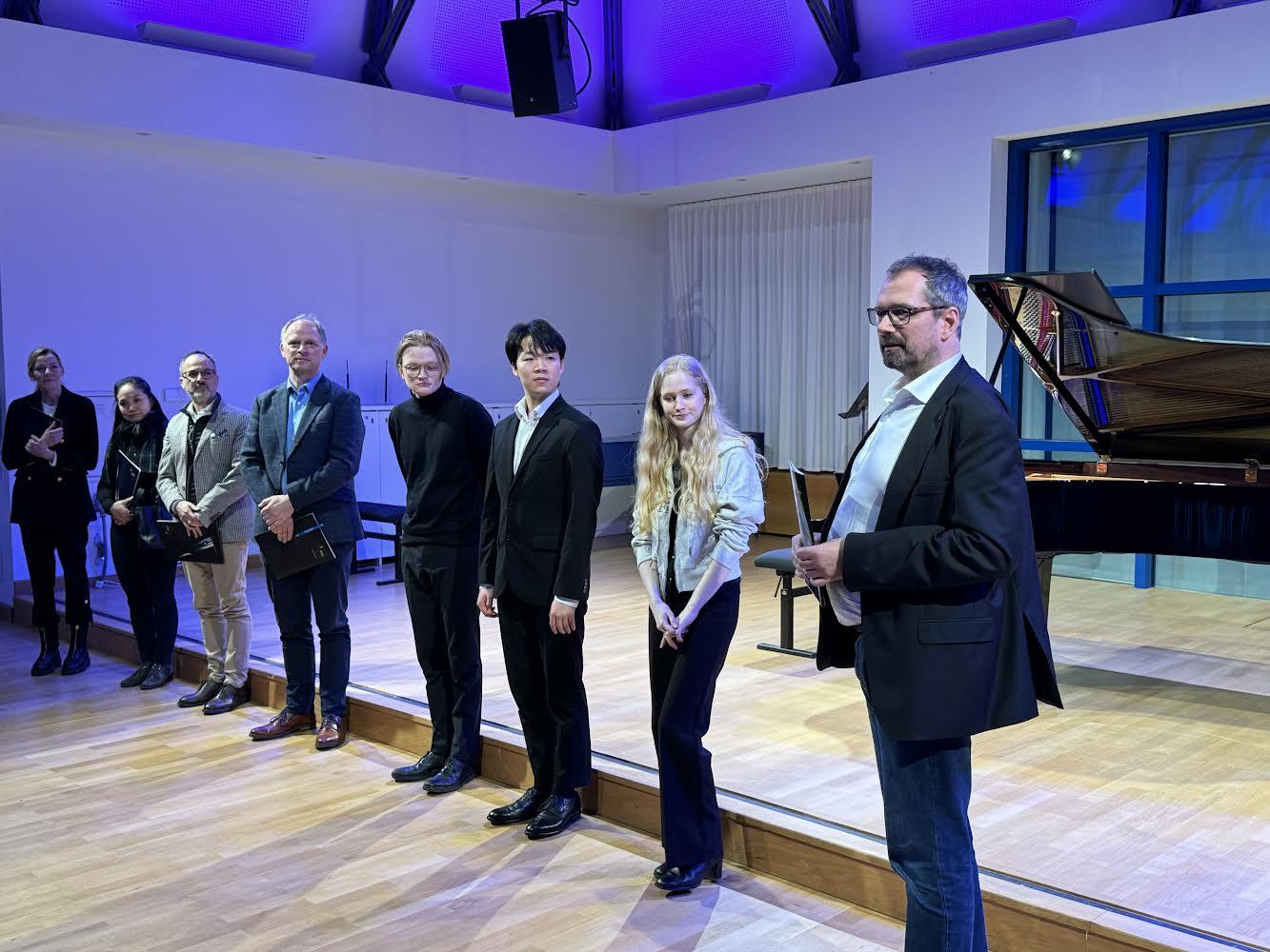 Die Stipendienvergabe beim Finale in Stuttgart: Studierende und Jury-Mitglieder stehen in einer Reihe vor einer kleinen Bühne mit Klavier. Die Personen schauen zu dem Mann ganz rechts, der gerade spricht.