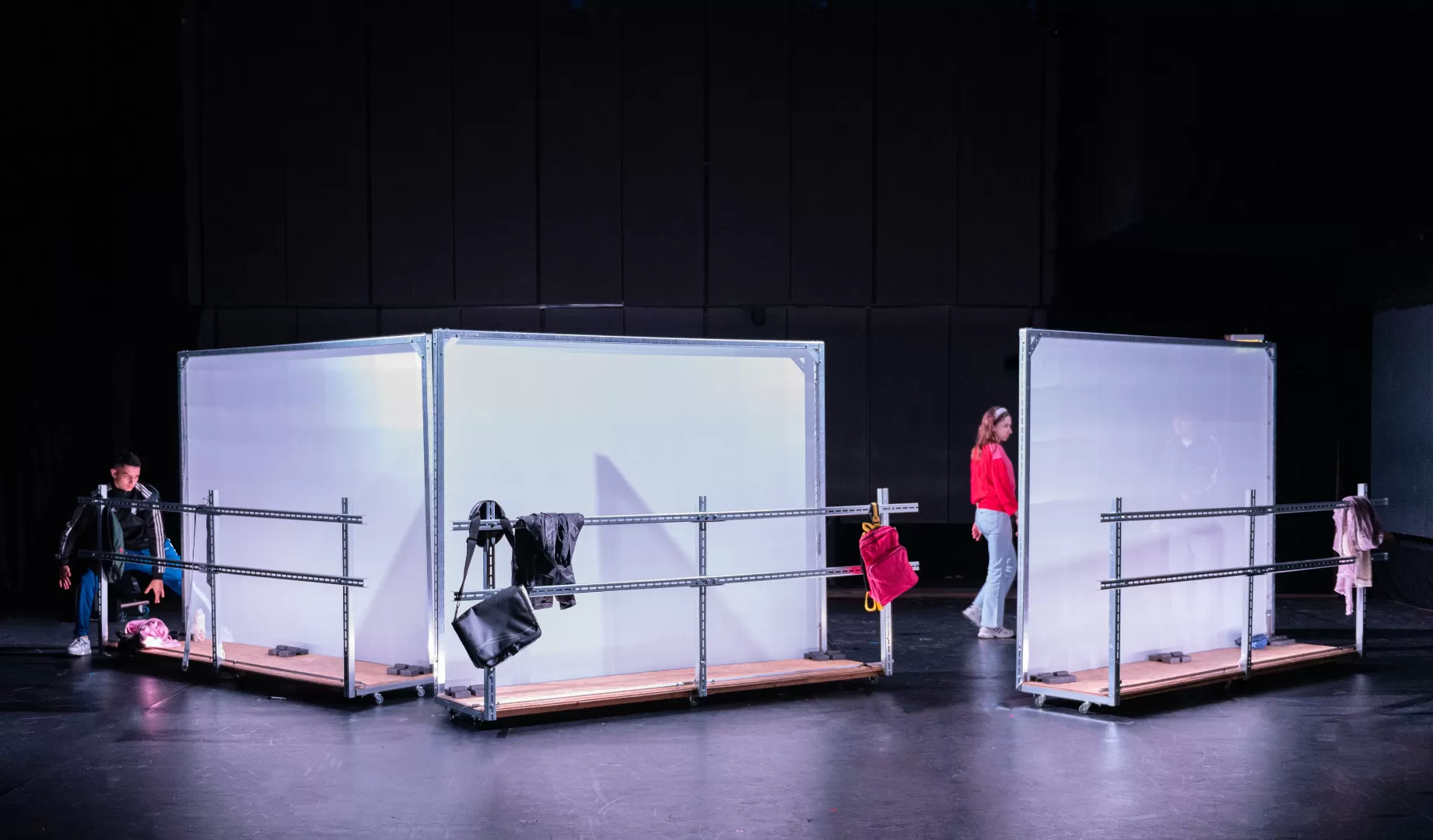 Drei einzelne Stellwände, weiß und leicht transparent, mit jeweils einer Metallbrüstung davor sind schräg gegeneinaner angeordnet, am linken Rand ist ein Mann zu sehen, in der Mitte eine Frau. An den Metallbrütungen hängen Kleidungsstücke und Taschen.