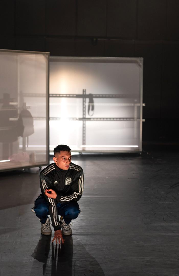 Ein Ausschnitt eines Bühnenraumes, im Hintergrund zwei weiße, halbtransparente Stellwände mit Metallbrüstung davor. Im Vordergrund hockt ein Mann in Turnschuhen und Adidas Trainingsanzug, er schaut fragend nach rechts.