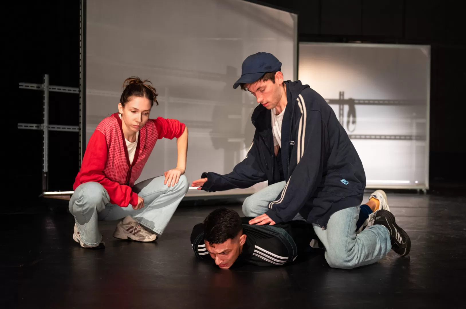 Auf dem Bild sind drei Personen in einem Bühnenraum zu sehen. Rechts zwei Männer, einer davon liegt bäuchlings am Boden, der andere kniet mit einem Knie auf ihm und drückt ihn nieder. Links daneben hockt eine Frau in Jeans, Turnschuhen und roter Jacke, die auf den am Boden liegenden Mann blickt.