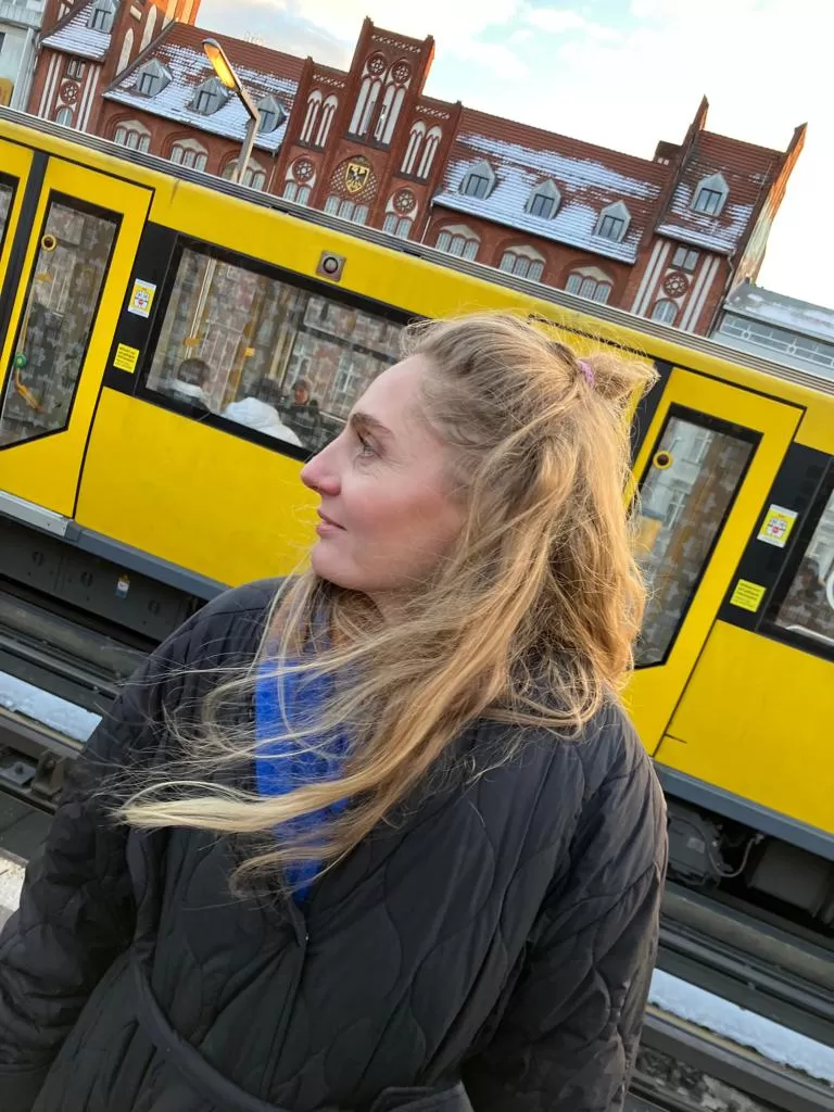 Eine Frau steht im Vordergrund, sie ist bis zur Hüfte zu sehen, trägt einen schwarzen Daunenmantel und hat lange, rotblonde Haare. Sie blickt nach links aus dem Bild. Hinter ihr ist eine gelbe Straßenbahn zu sehen, dahinter im oberen Teil des Fotos ist ein Teil des Gebäudes Altes Postamt in Berlin-Kreuzberg zu sehen.