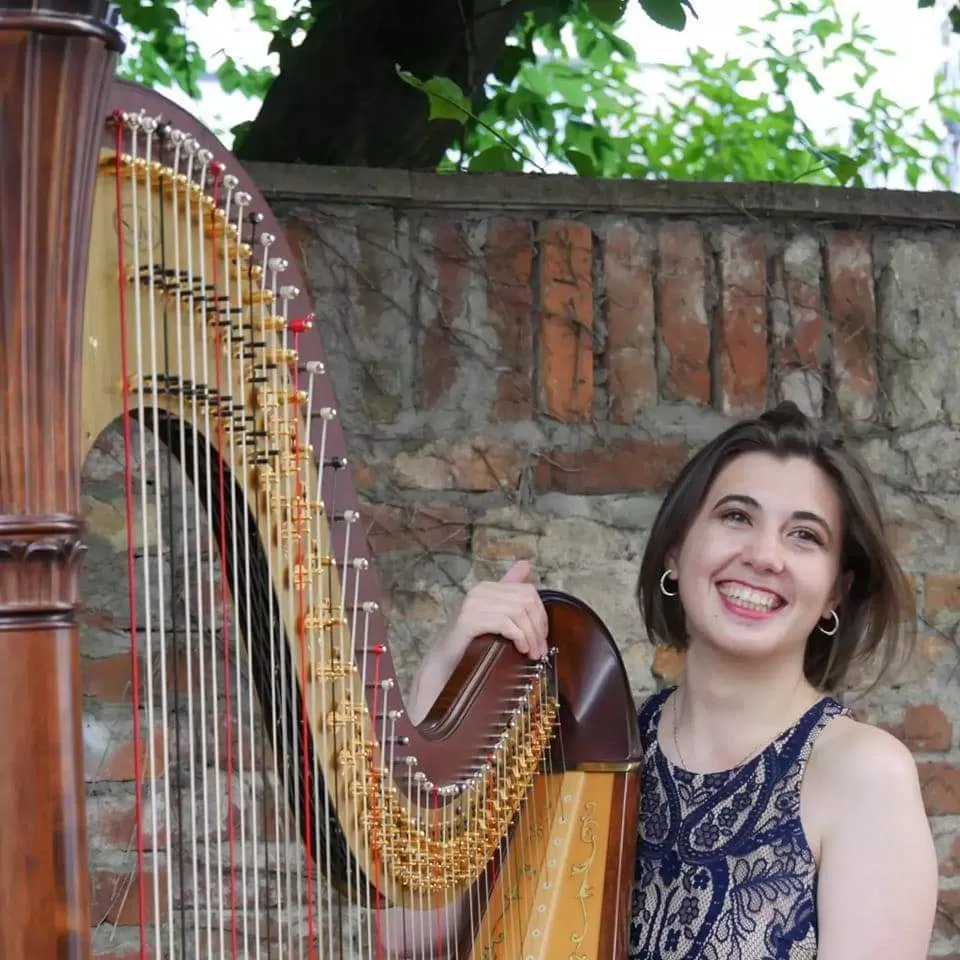 Gwenaëlle Le Meignen sitzt an ihrer Harfe und lacht. Sie hat schultelange, braune Haare und trägt ein Kleid mit blau-beigem Muster. Hinter ihr ist eine Ziegelsteinmauer zu sehen, dahinter ein Baum.