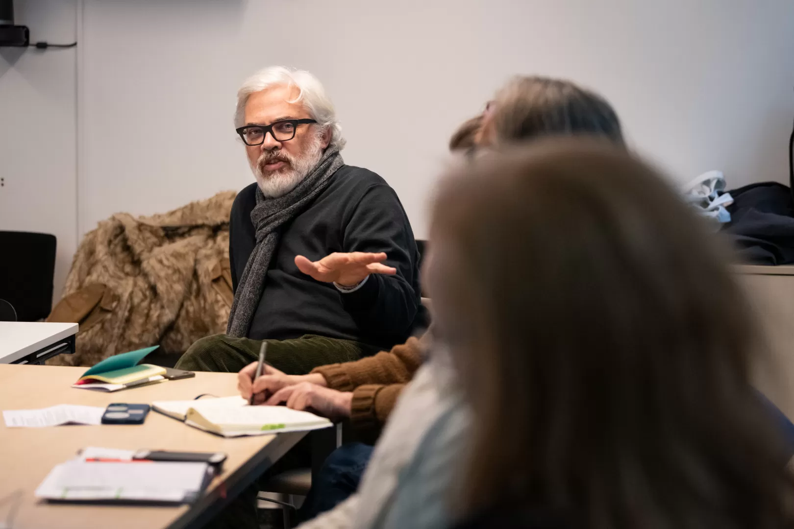 Drei Personen sitzen an Tischen, vor ihnen Dokumente. Zwei Personen im Vordergrund sind nicht gut erkennbar, aber der Mann mittig im Bildhintergrund ist im Fokus. Er hebt die linke Hand, um etwas zu erklären.