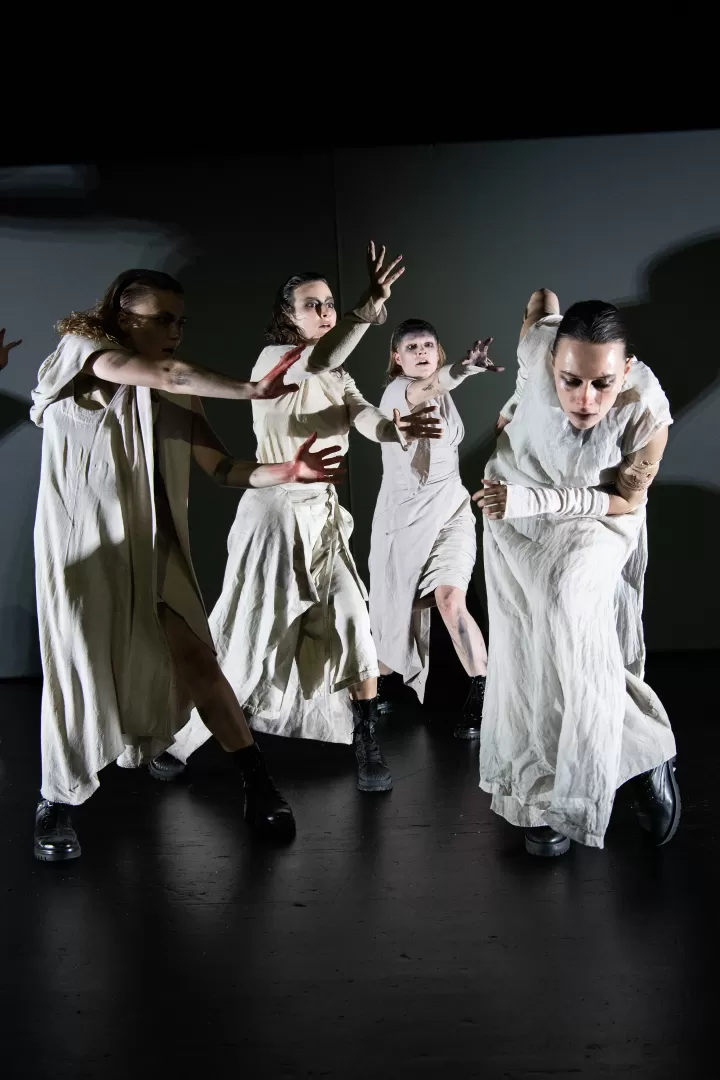 Vier Performer*innen in weißen Toga-Gewändern bewegen sich schnell und dynamisch auf der Bühne