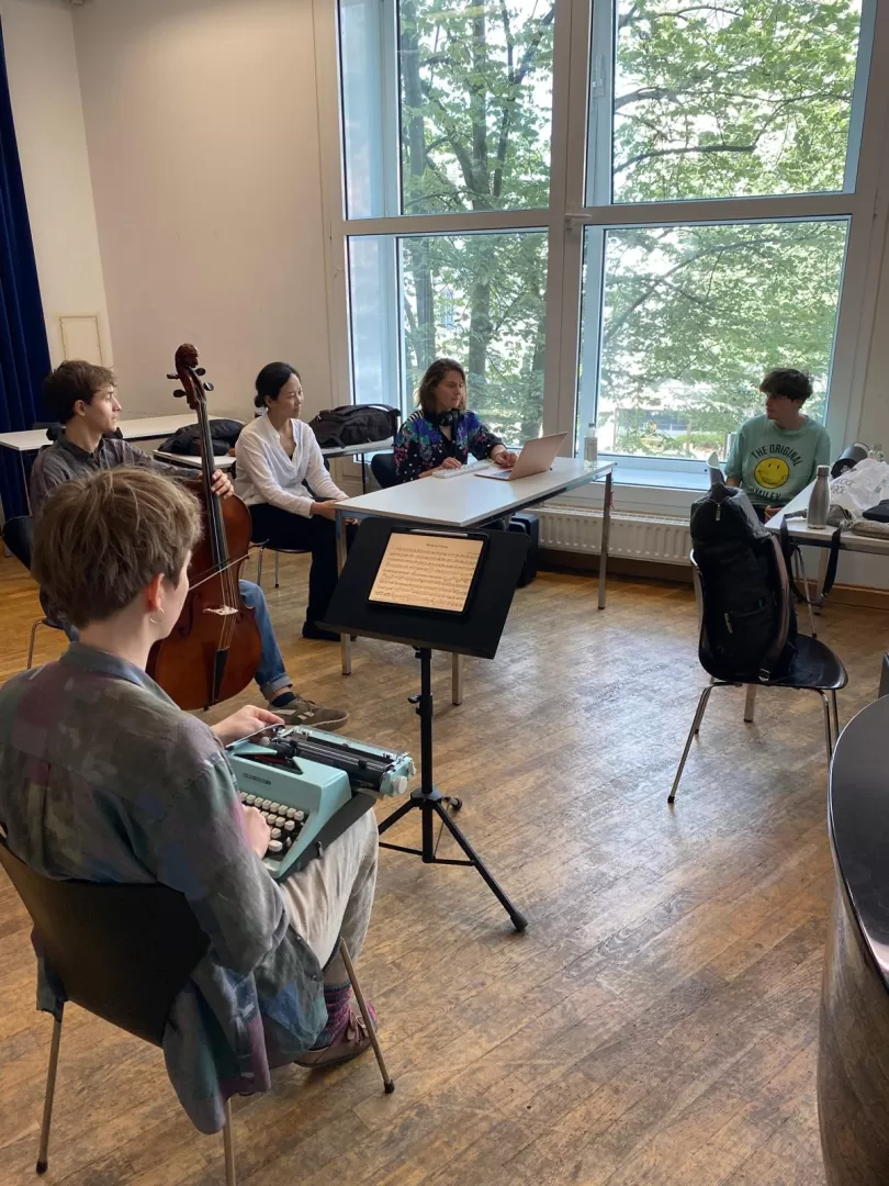 Fünf Studierende sitzen in einer Probe zusammen.  Eine Studentin mit kurzen braunen Haaren sitzt links mit dem Rücken zur Kamera im Bildvordergrund. Sie hat eine Schreibmaschine auf dem Schoß. Hinter ihr sitzt ein Student mit einem Cello. Die drei übrigen Studierenden sitzen an Tischen. Sie unterhalten sich.