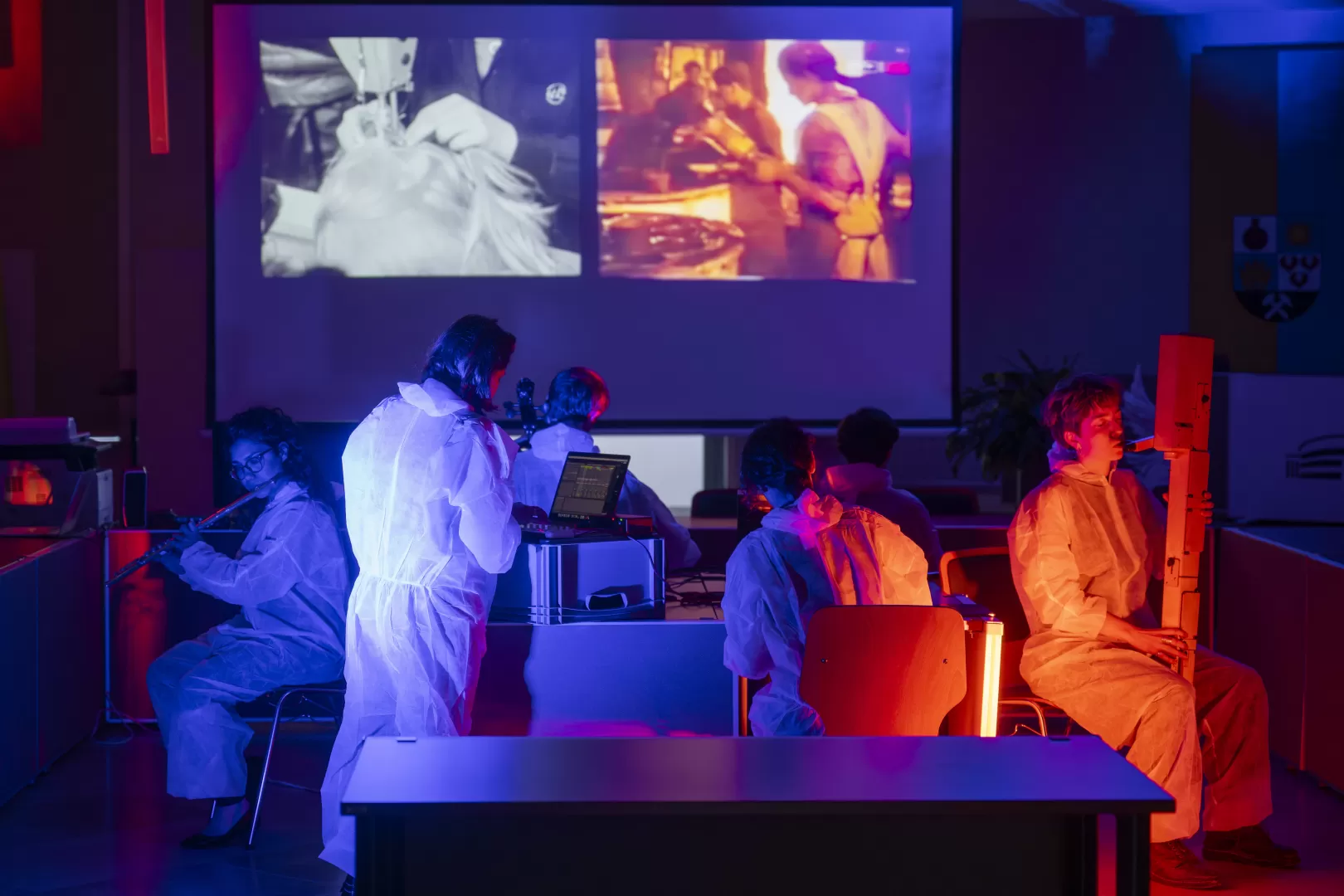 Eine Gruppe Musiker*innen sitzt mit verschiedenen Instrumenten um einen Tisch herum und musiziert. Alle tragen weiße Einweg-Overalls. Eine Studentin links im Bild steht an einem Laptop. Im Hintergrund sieht man historische Fotos, die an die Wand projiziert werden. Der Raum ist in blaues und rot-orangenes Licht getaucht.