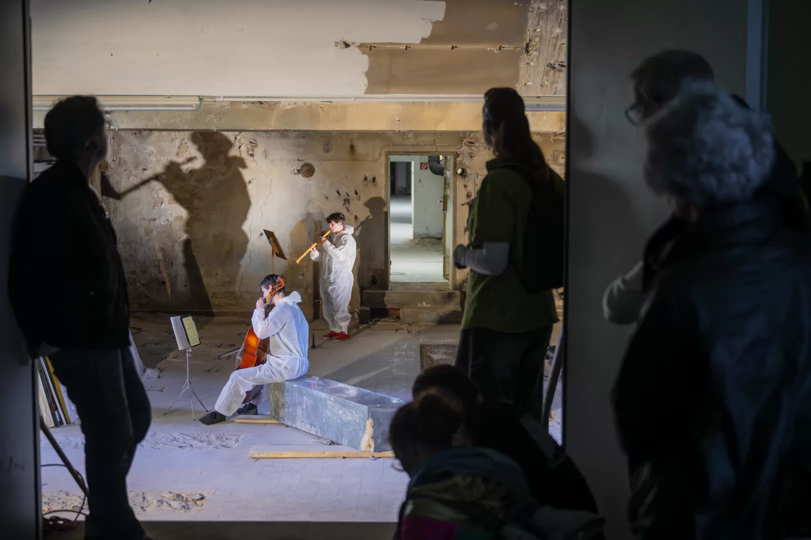 In einem Kellerraum musizieren zwei Studierende mit Cello und Oboe. Sie tragen weiße Einweg-Overalls. Durch das Licht werden ihre Schatten groß an die Wand geworfen. Im Vordergrund des Bilds sind einige Zuschauer*innen zu sehen, die durch eine Türöffnung in den Raum schauen.