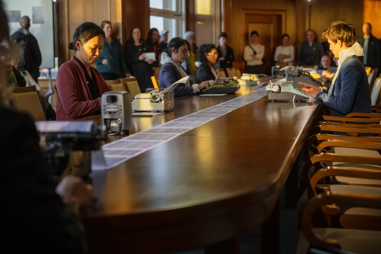 In einem Raum mit Holzvertäfelung sitzen die Studierenden an einem langen Holztisch verteilt und tippen auf Schreibmaschinen. Sie haben nun Alltagskleidung über ihre Overalls gezogen. Das Publikum steht am Rand und schaut zu.