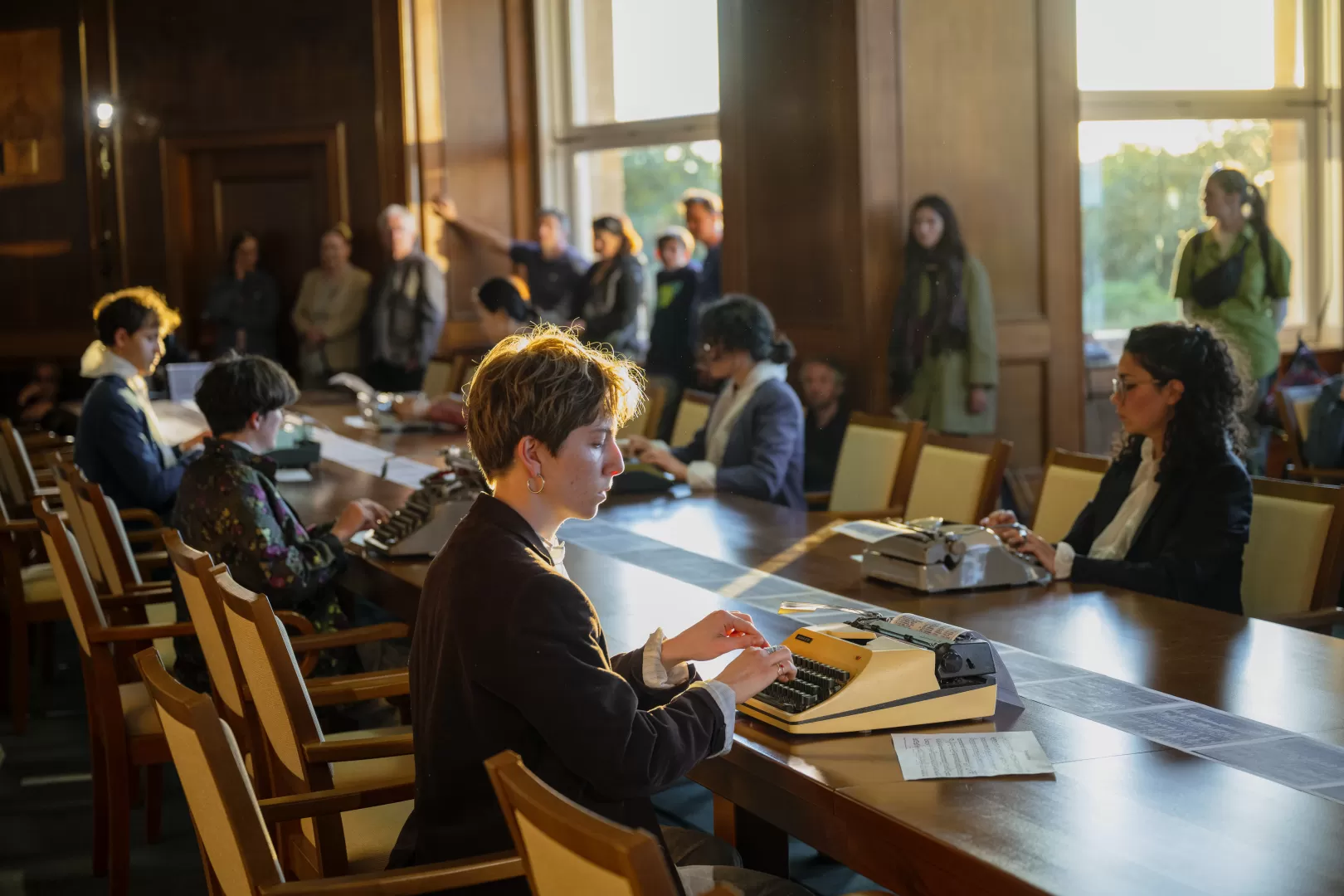 In einem Raum mit Holzvertäfelung sitzen die Studierenden an einem langen Holztisch verteilt und tippen auf Schreibmaschinen. Sie haben nun Alltagskleidung über ihre Overalls gezogen. Im Hintergrund sind große Fenster zu sehen. Das Publikum steht am Rand und schaut zu. Eine Studentin mit kurzen braunen Haaren und einem schwarzen Blazer ist hier im Vordergrund.