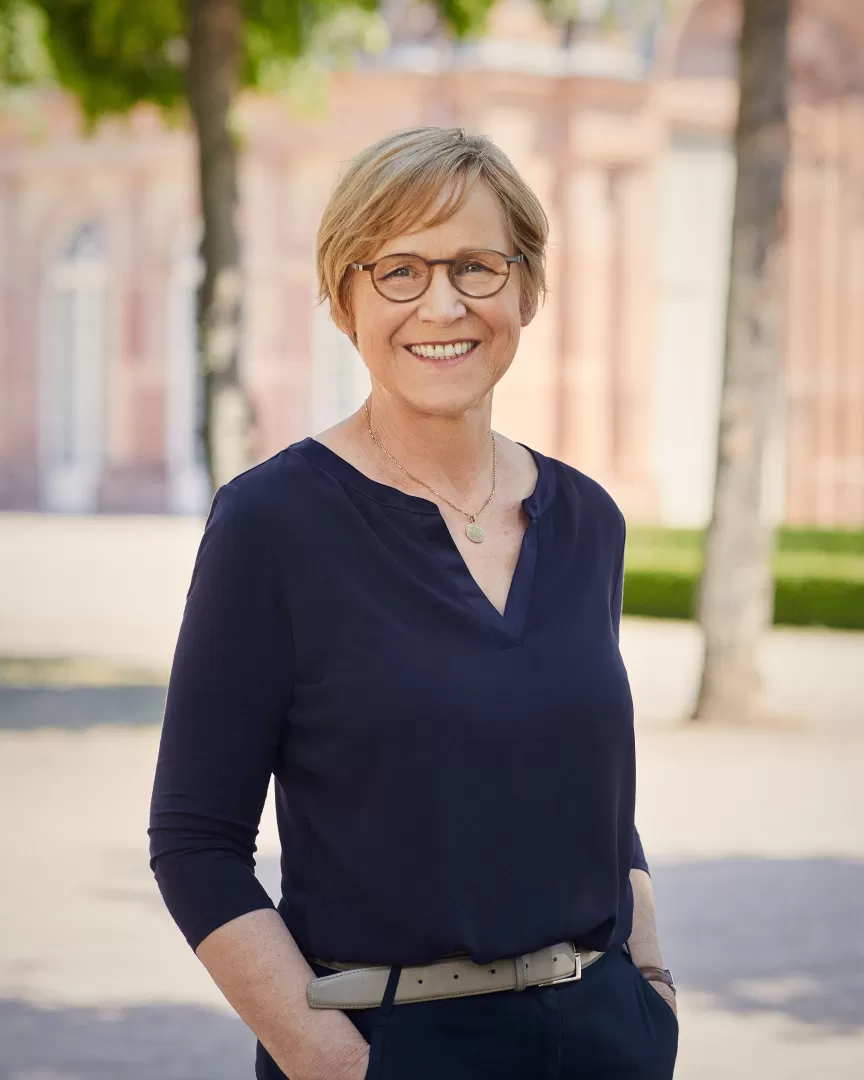 Portraitfoto von Heike Hoffmann. Sie hat kurze blonde Haare, trägt eine Brille und lächelt in die Kamera. Ihre Hände hat sie in den Hosentaschen. Sie trägt eine blaue Bluse, eine blaue Hose und eine goldene Kette.