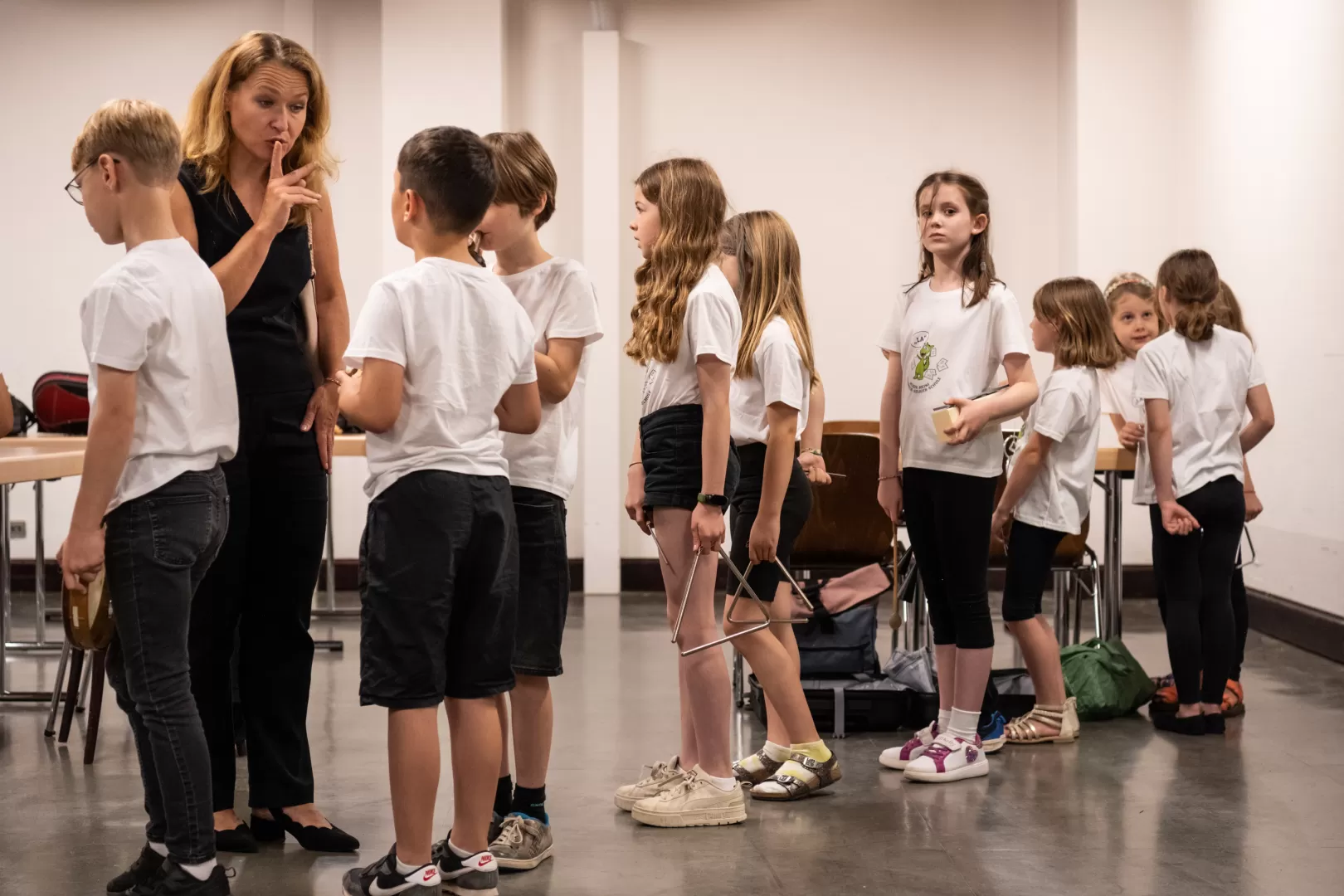 Hinter den Kulissen hat sich eine Grundschulklasse in weißen Shirts für den Auftritt bereit gemacht und steht in einer Schlange. Die blonde Lehrerin (links im Bild) legt ihren Finger auf die Lippen und bedeutet damit einem Schüler, ruhig zu sein.