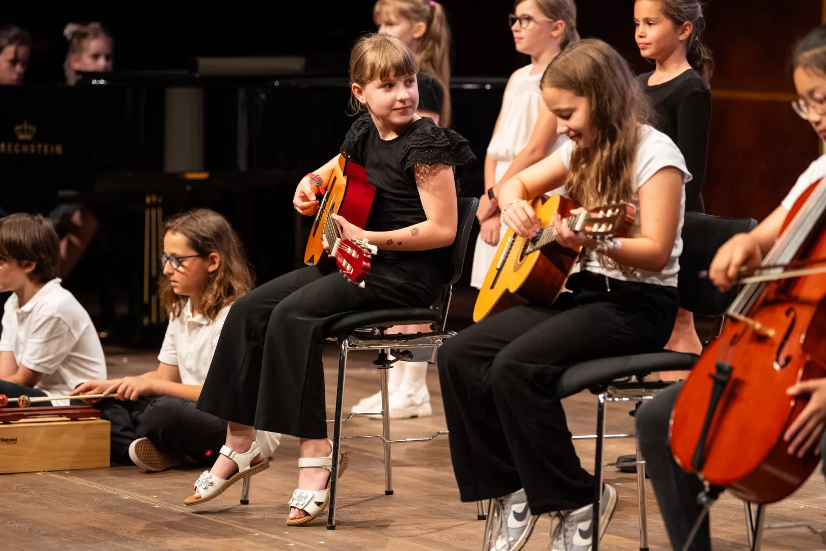 Eine Gruppe Kinder sitzt mit Instrumenten auf der Bühne. Im Fokus stehen zwei Mädchen, die auf Stühlen sitzen und Gitarre spielen. Um sie herum sitzen weitere Kinder, links auf dem Boden eines mit Xylophon, rechts auf einem Stuhl eines mit Cello.