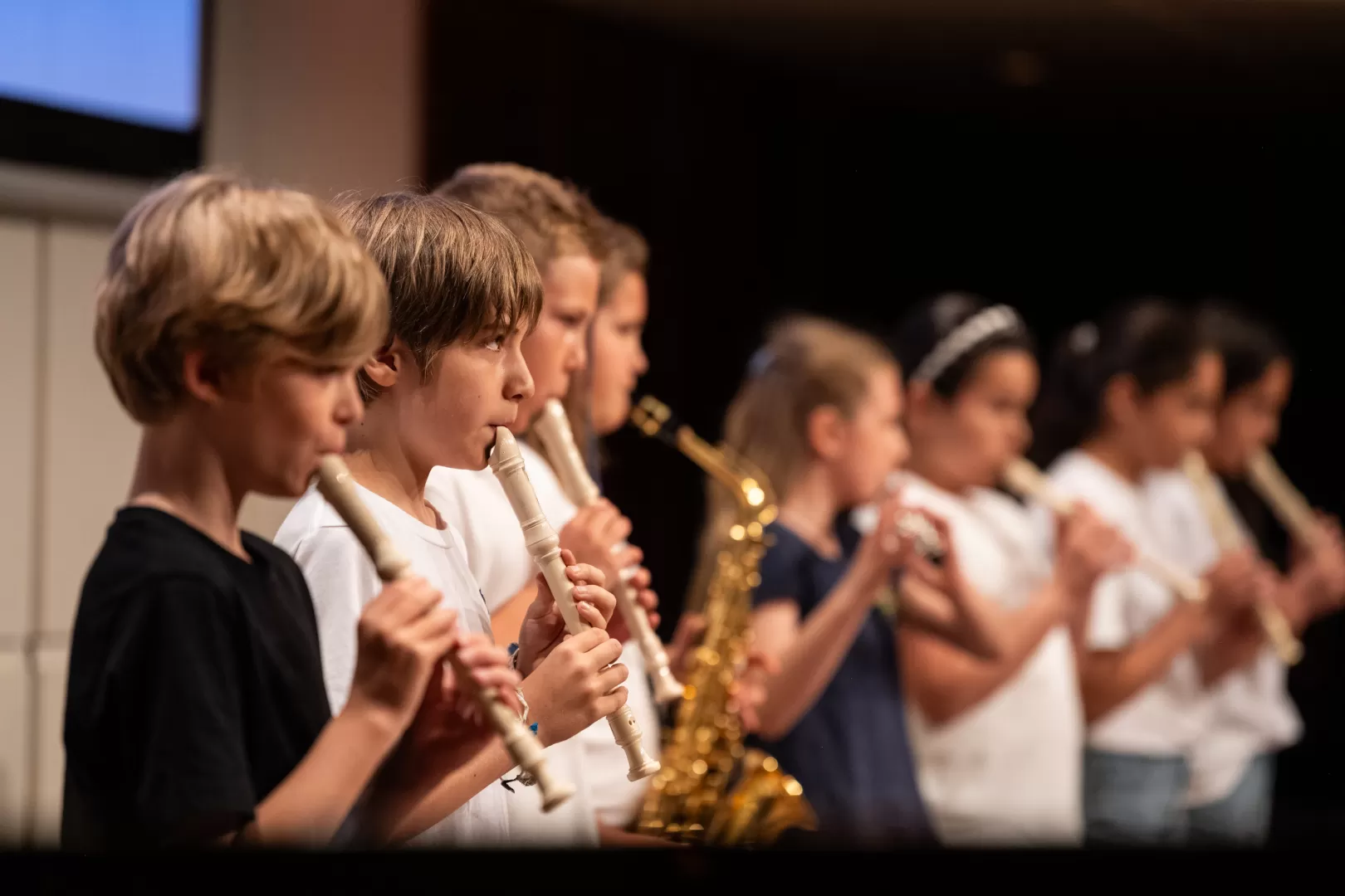 Drei Jungs spielen Blockflöte. Verschwommen im Hintergrund ist zu erkennen, dass weitere Kinder Saxophon und Flöte spielen.