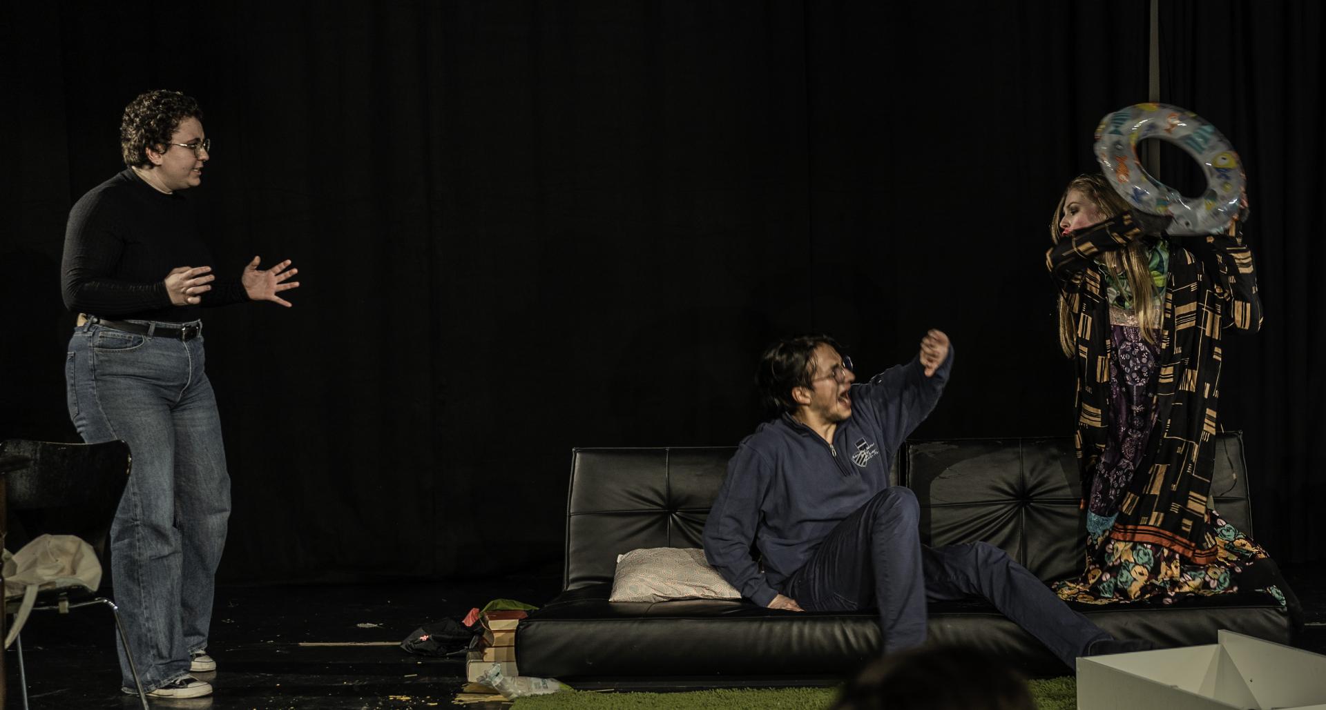 ein junger Mann sitzt auf einem Sofa rechts im Bild. Eine junge Frau in bunter schriller Kleidung hält einen Schwimmreifen hoch und bedroht den Mann damit. In der linken ecke steht eine andere junge Frau und erhebt die Hände, wie als würde sie einschreiten wollen.