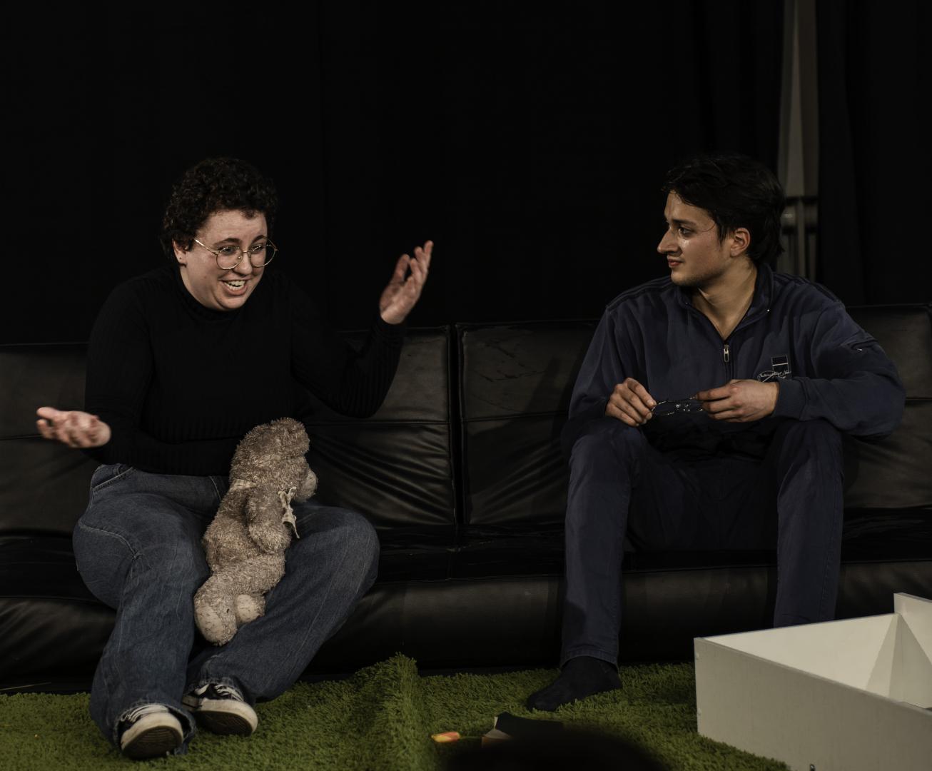 ein junger Mann und eine junge Frau sitzen auf einem Sofa. Die Frau hat einen Teddy auf dem Schoß und ihre Hände sind gehoben, als würde sie nicht weiter wissen/sich erklären müssen. Der Mann blickt sie an.