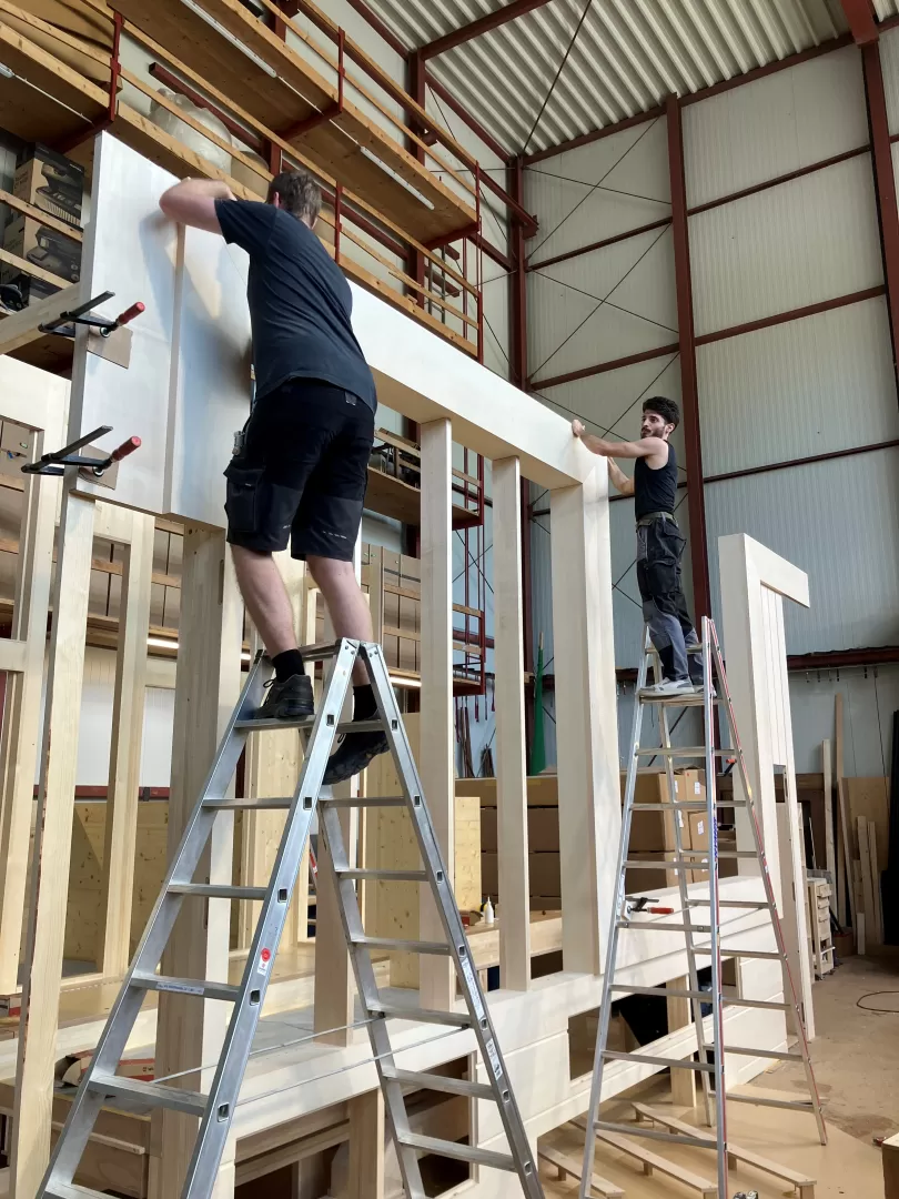 Zwei Männer stehen auf Leitern und arbeiten an einem großen Holzgehäuse.